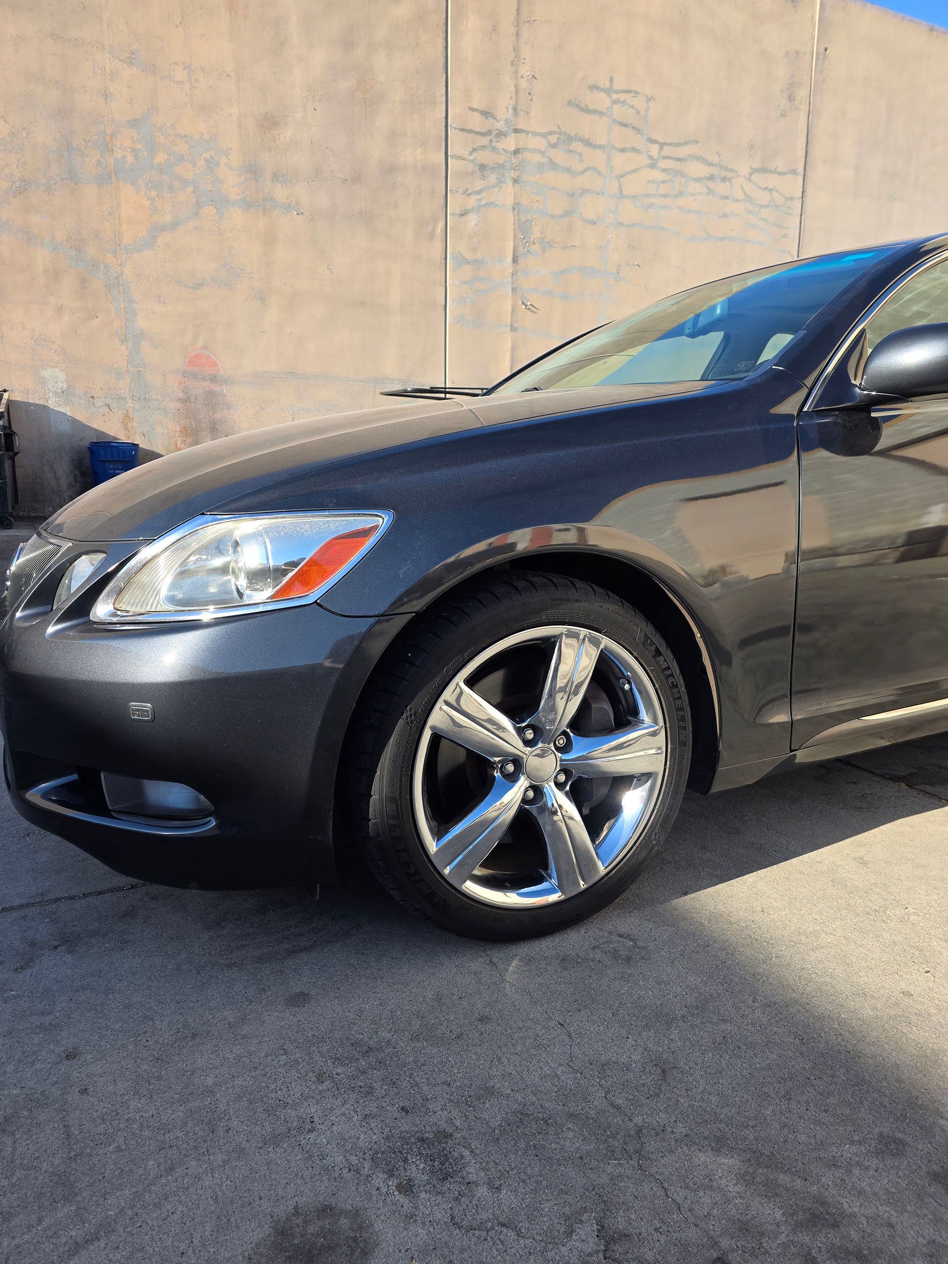 Dark gray sedan parked on pavement beside a concrete wall, front wheel and headlight visible