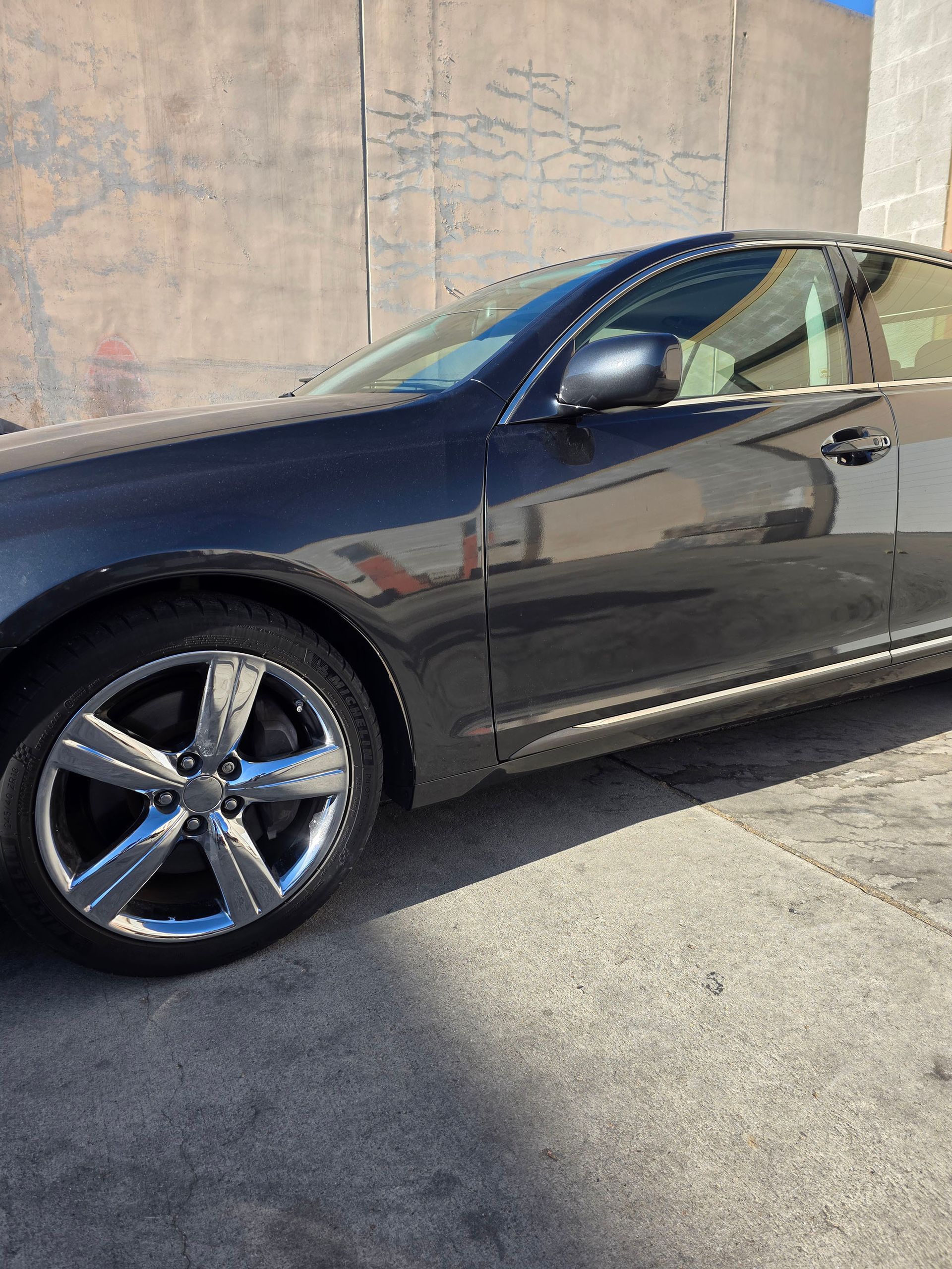 Dark gray coupe parked in sunlight beside a concrete wall, showing the front side and chrome wheel.