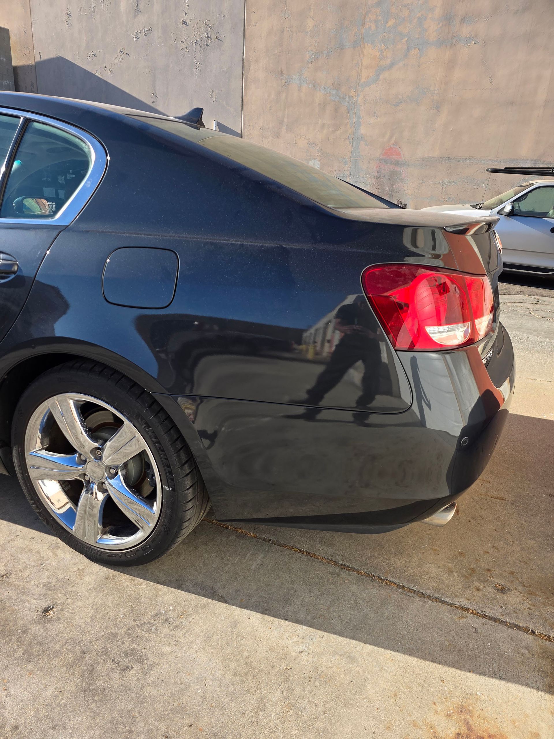 Black sedan parked on a sunlit concrete surface beside a wall, rear side view.