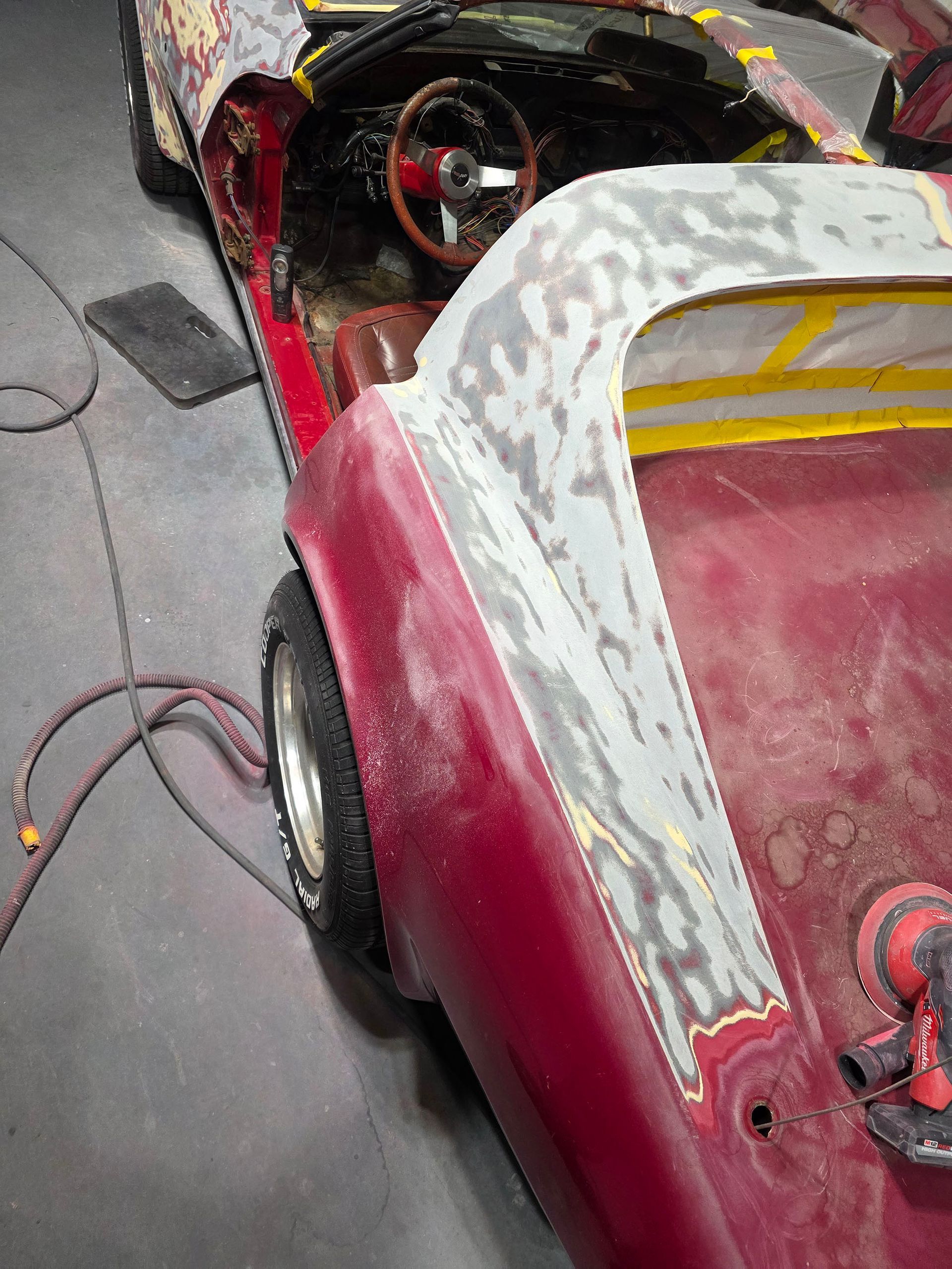Partially sanded red car body with exposed metal, seen in a garage with a steering wheel and cables nearby