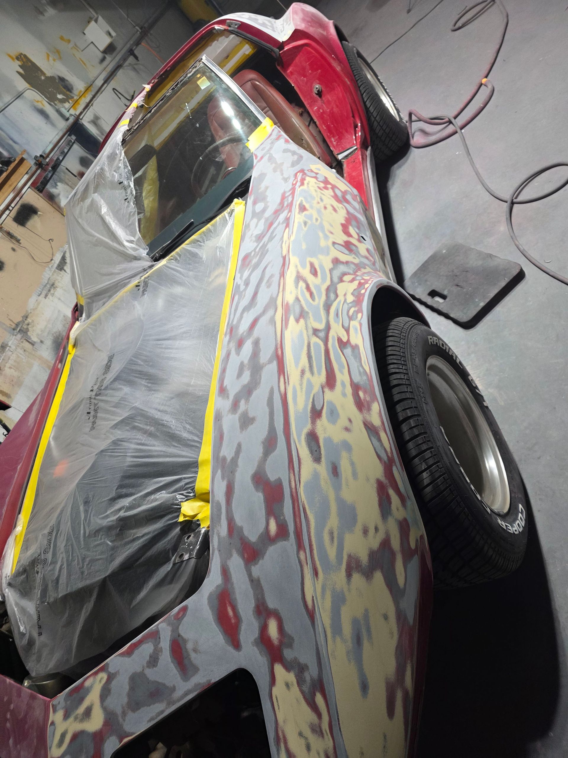 Damaged red car in a garage, covered with black plastic and gray primer patches, side view
