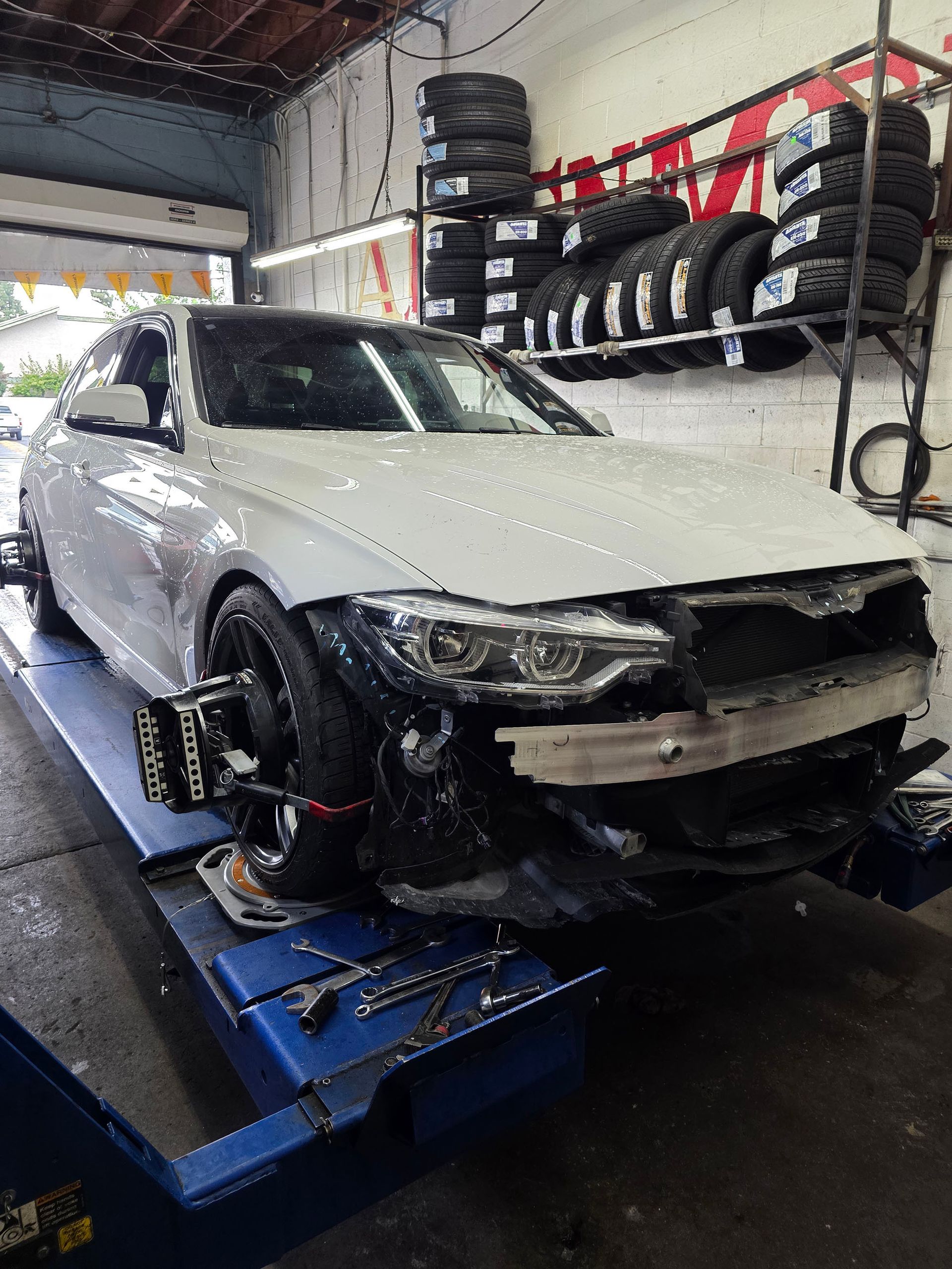 White sedan on a blue lift in an auto repair shop, with front-end damage and missing bumper.
