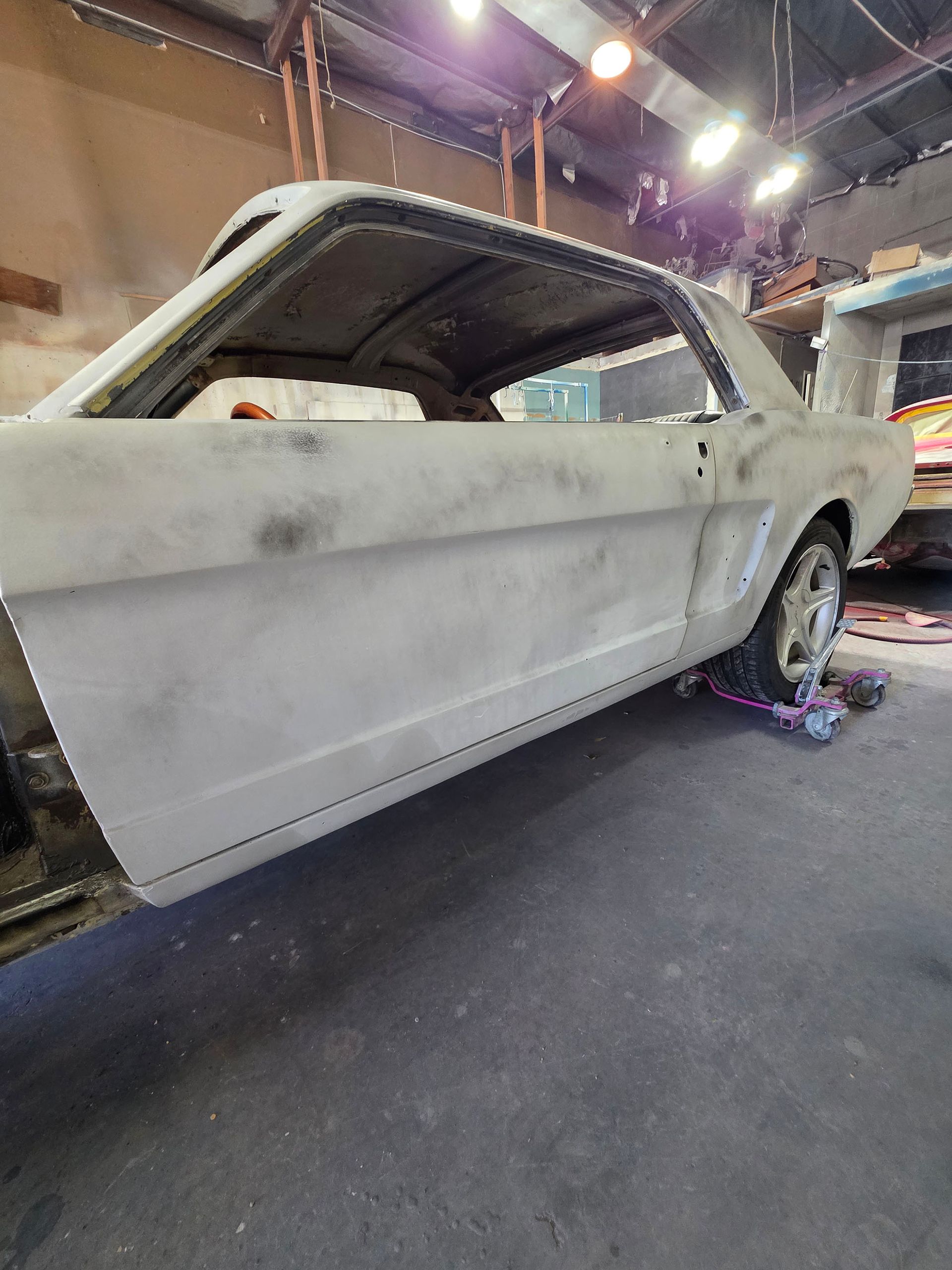 Primered classic car body in an auto shop, with wheels on and workshop lights overhead.