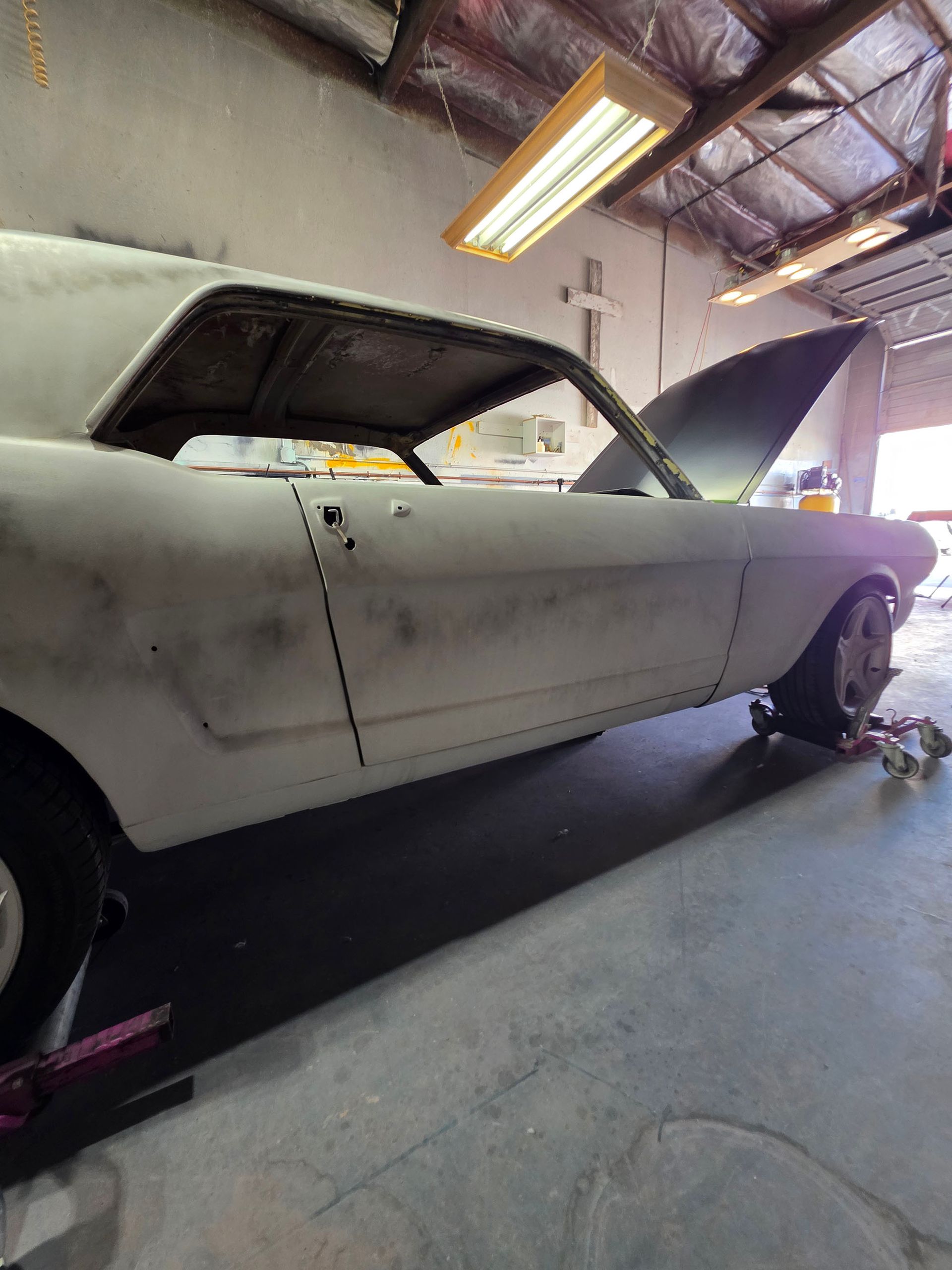 White classic car in a garage with its hood open and front wheel removed, under fluorescent lights.