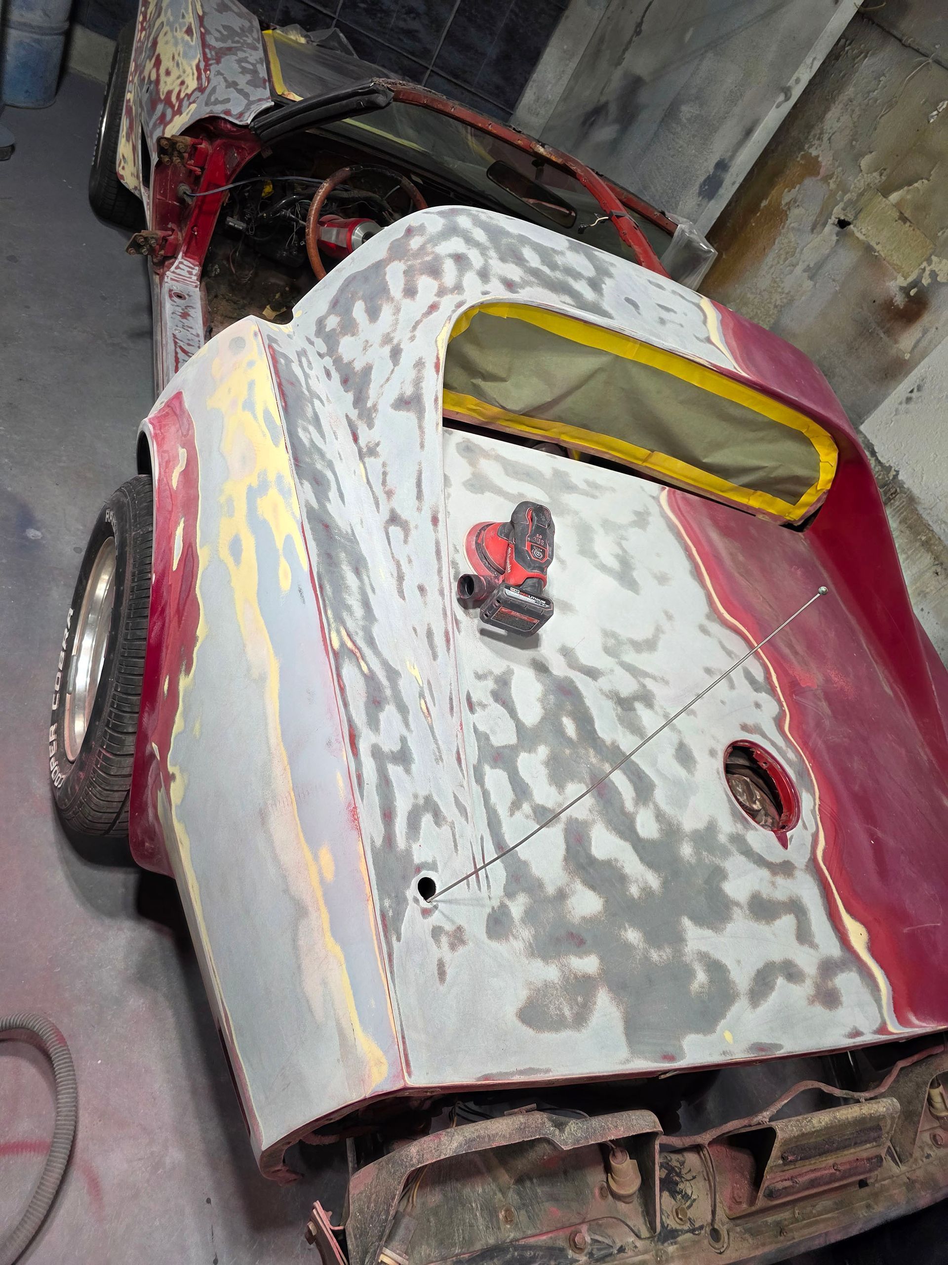 Rusty red vintage car body with a yellow rear window, parked indoors among tools and debris