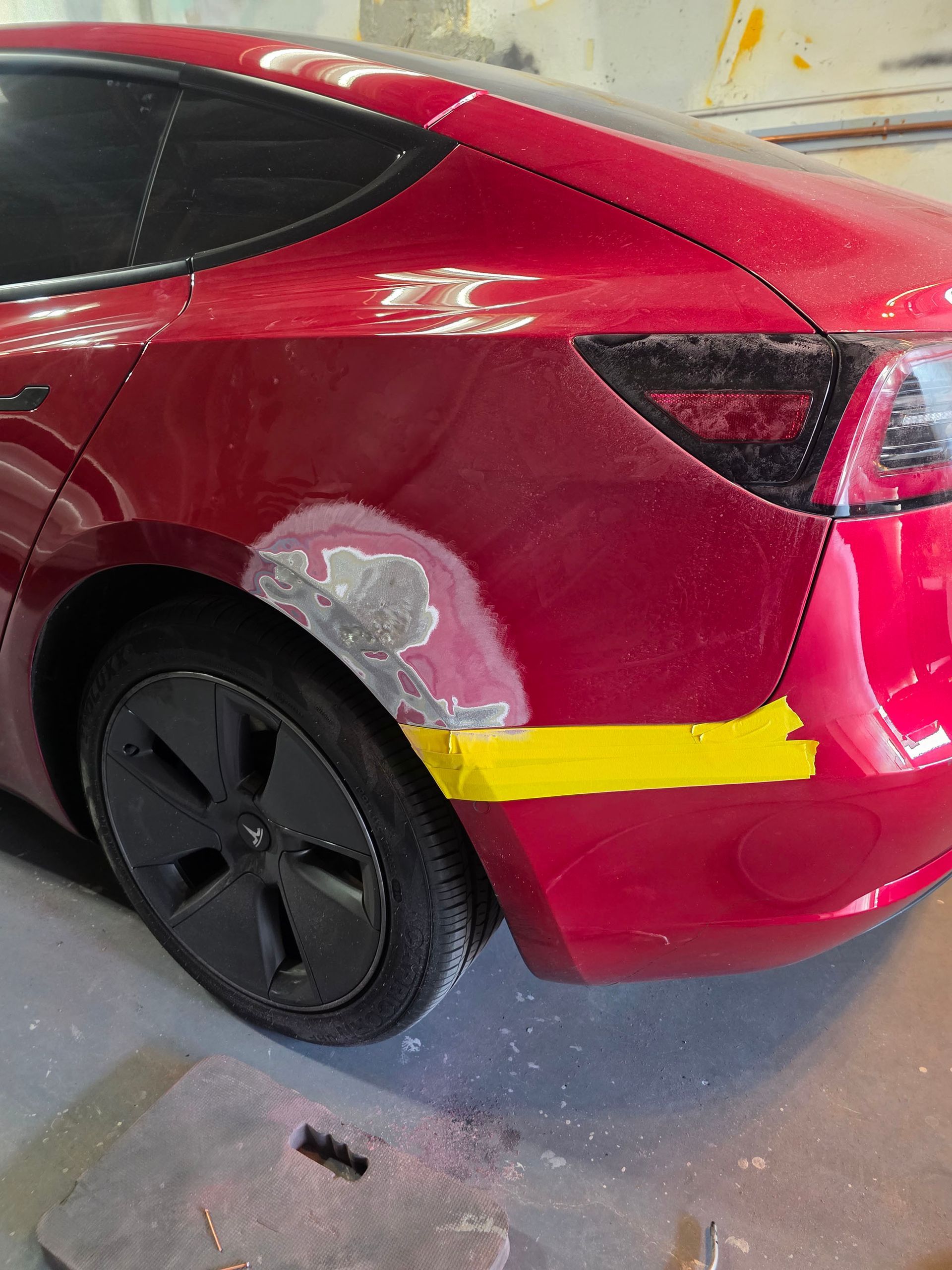 Red car’s rear quarter panel with gray body filler and a yellow paint strip in a garage.