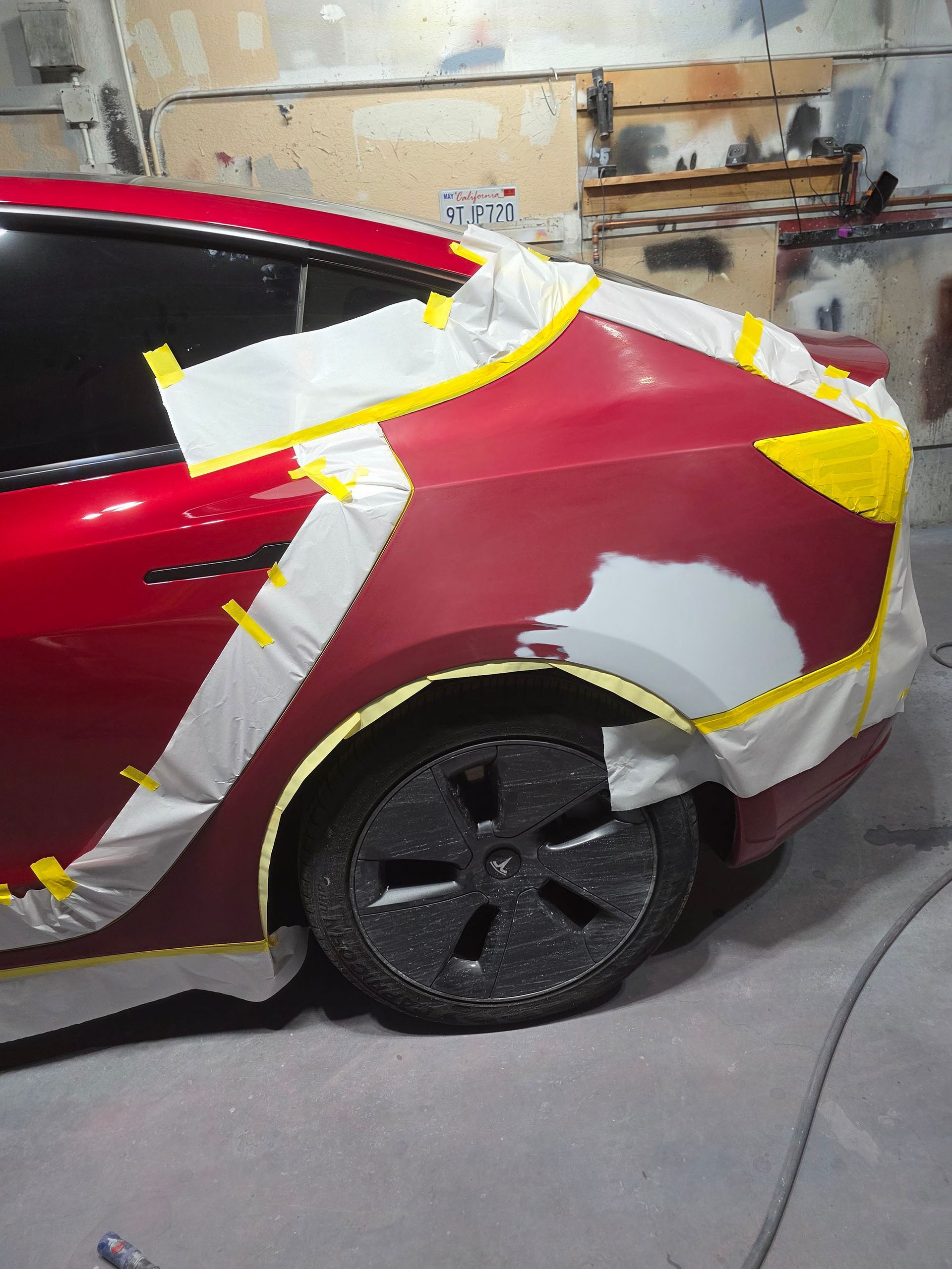 Red car body with white and yellow masking tape in a garage, mid-paint or repair.