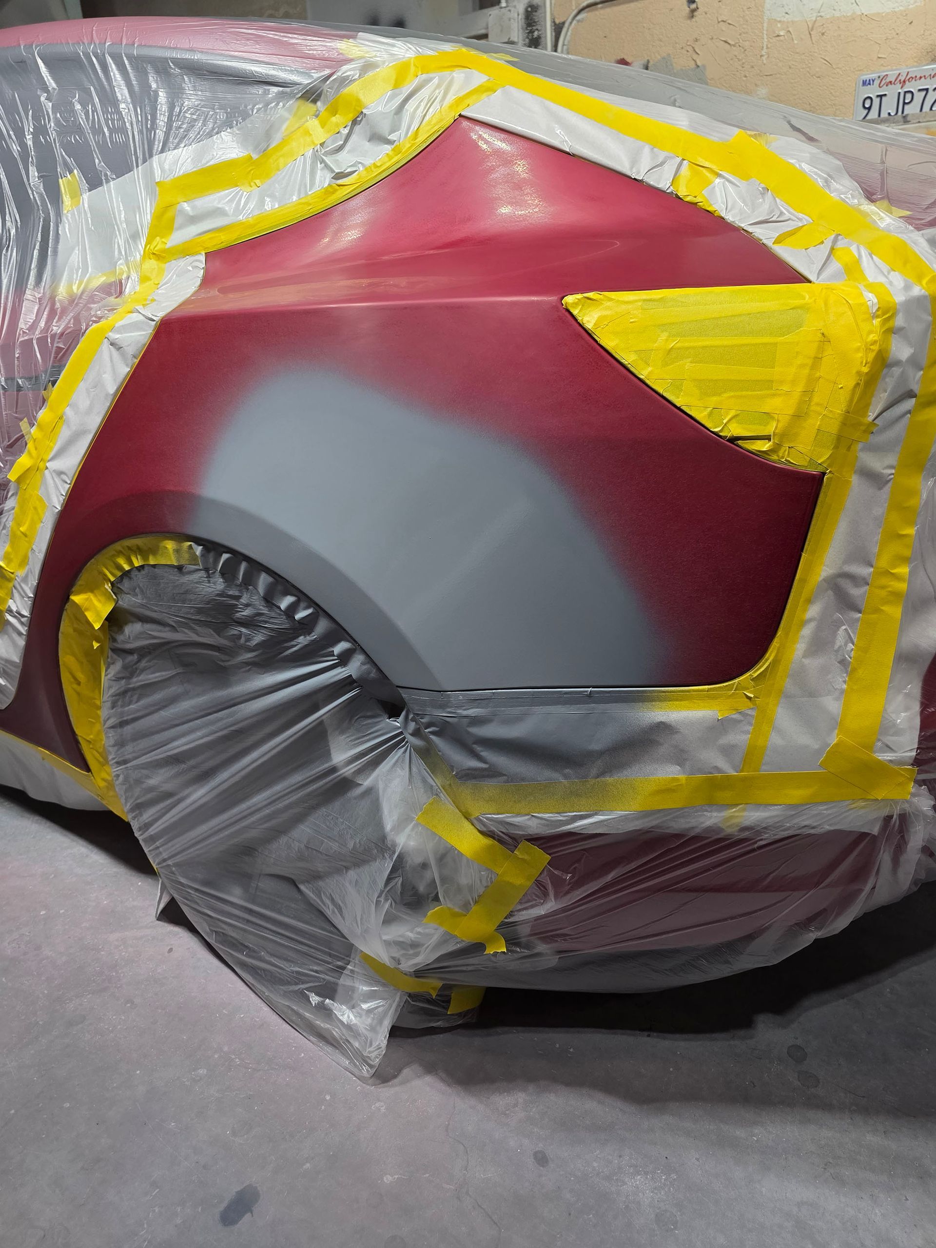 Red car body panel with gray primer, partially masked with tape and plastic in a workshop