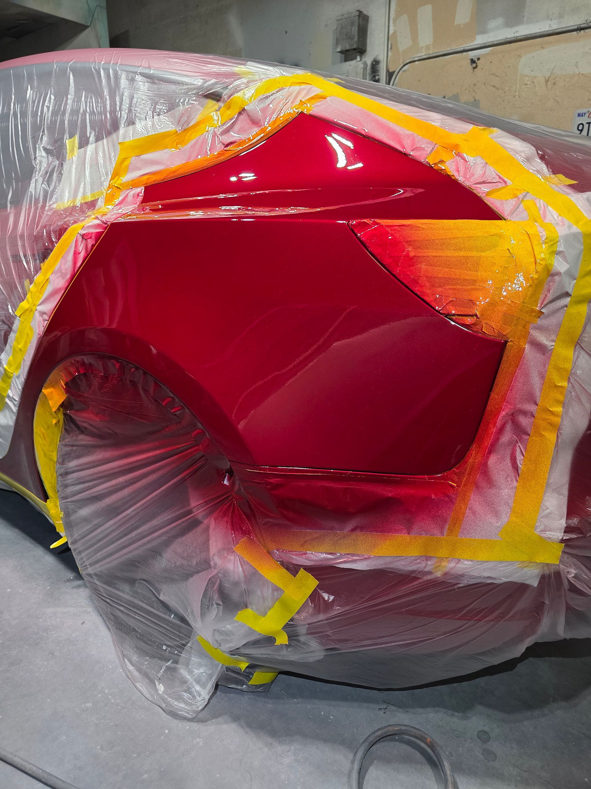 Red car front end wrapped in clear plastic, with red and yellow paint or masking around the headlight area