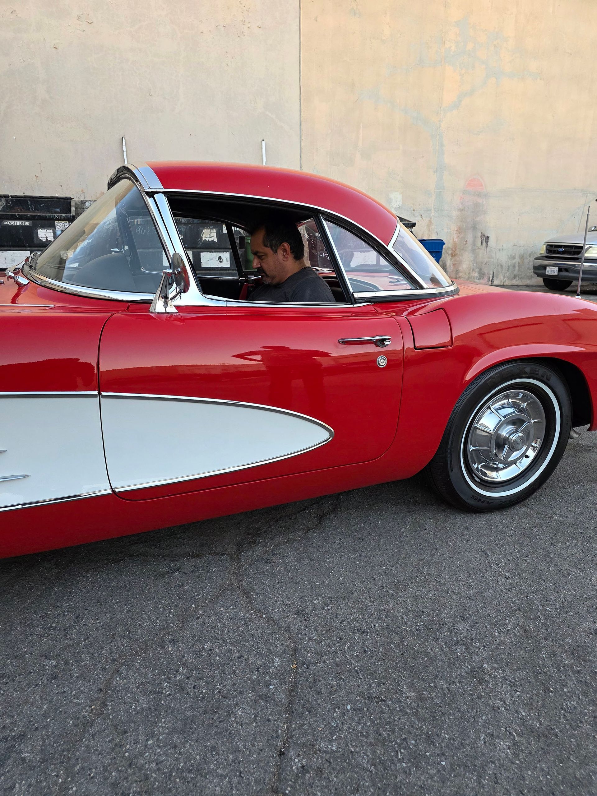 Red classic car with white side panel parked on pavement, driver visible inside against a beige wall.