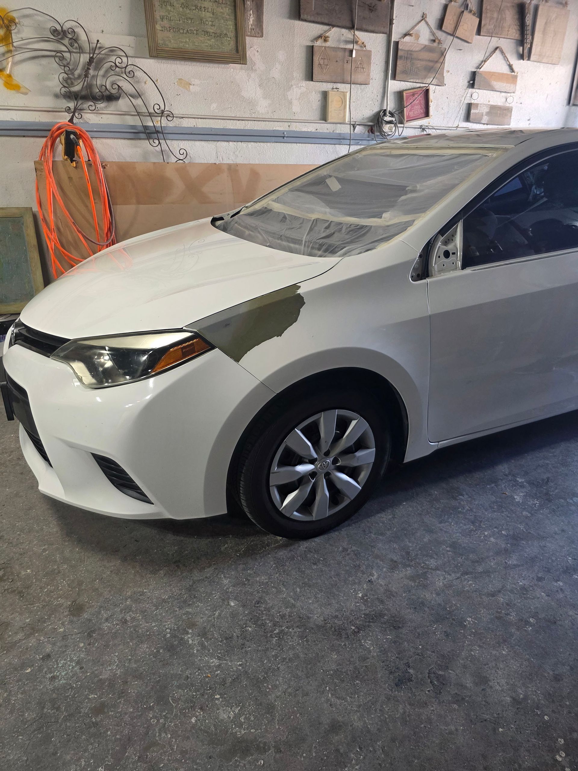 White sedan parked in a garage, front-left view with dark tape on the hood and fender.