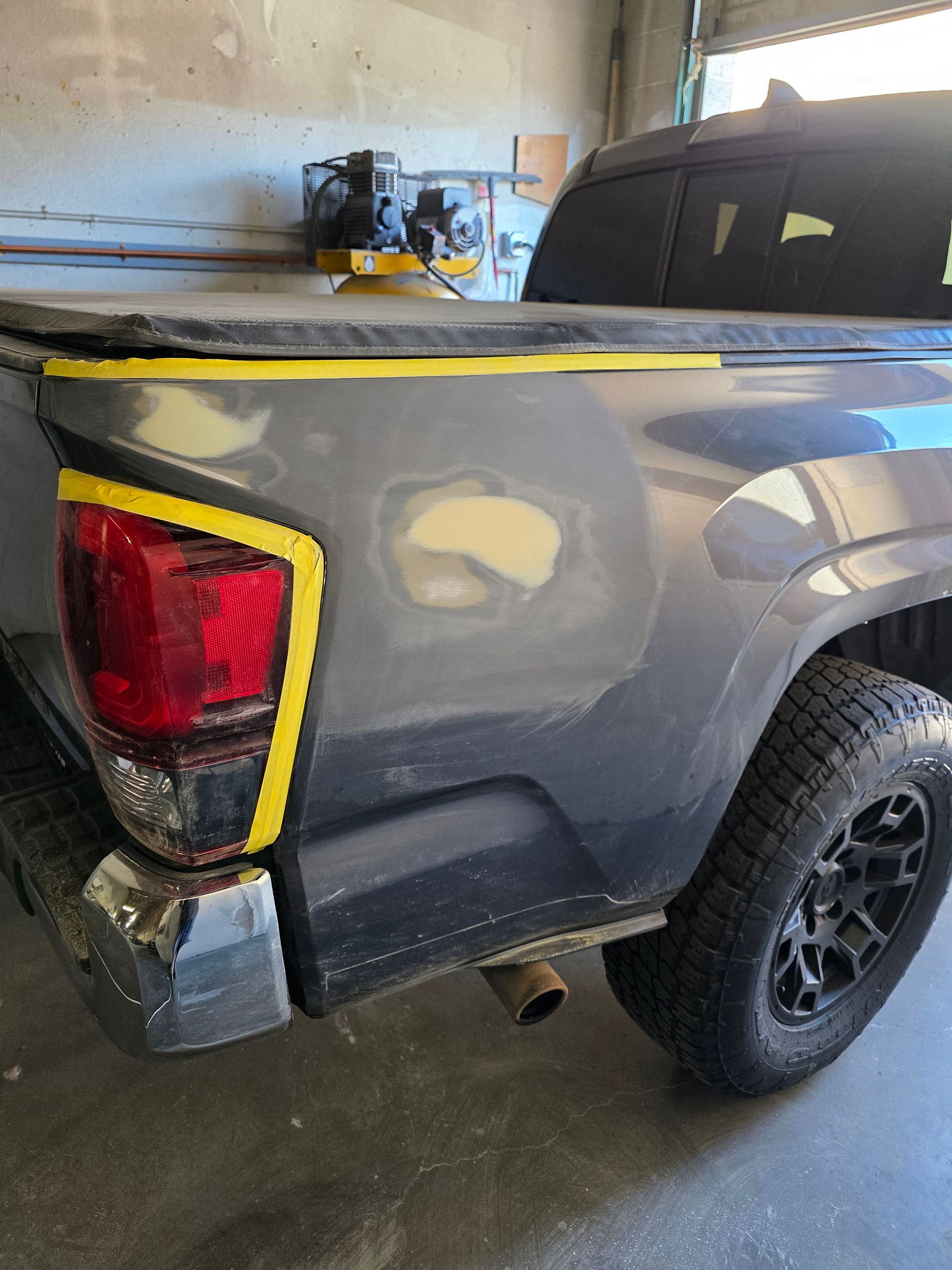 Gray pickup truck with yellow masking tape and body filler patches in a garage