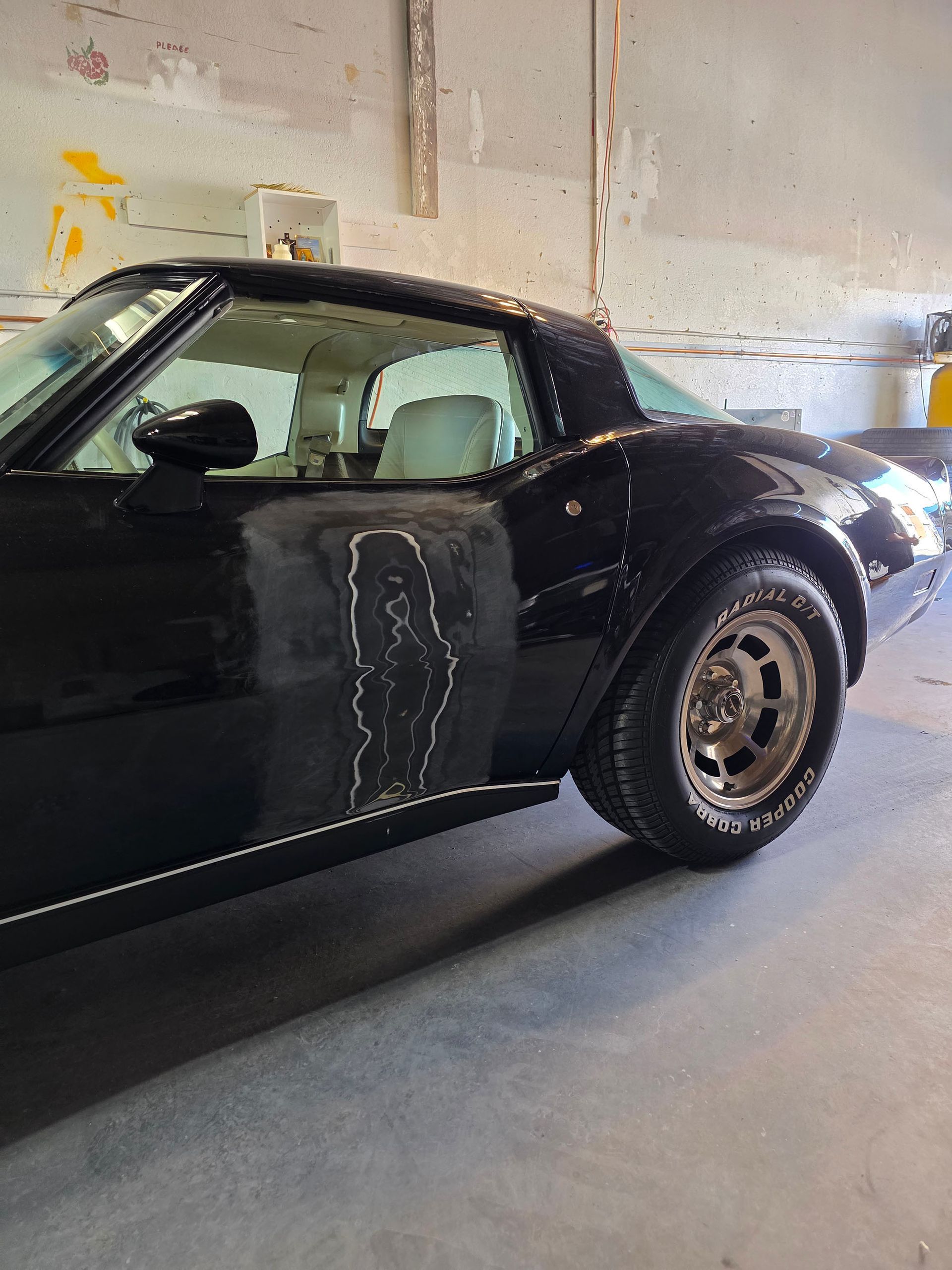 Black classic coupe parked indoors in a garage, side view showing the driver door and rear wheel.
