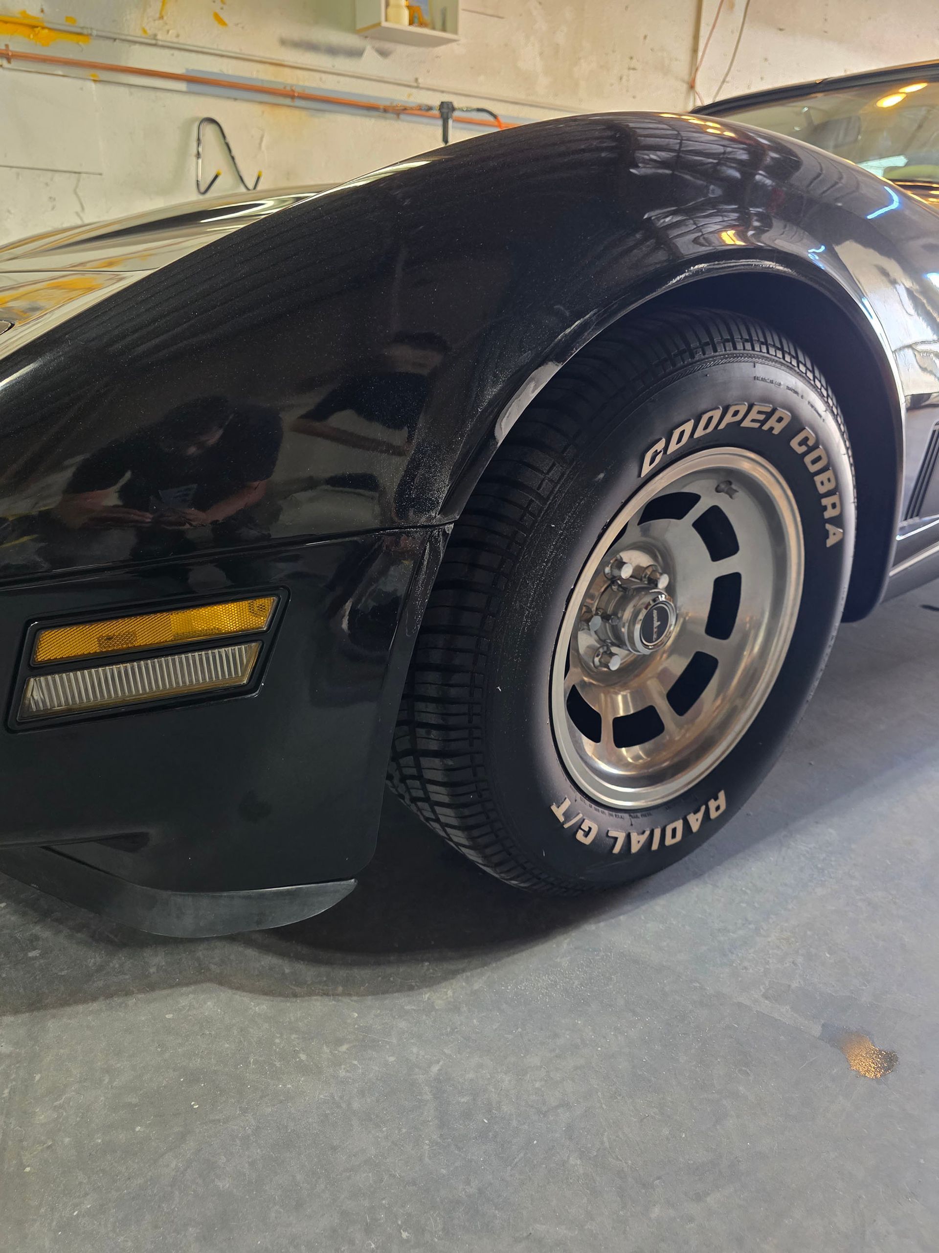 Black classic sports car front wheel and fender in a garage, with chrome rim and white-letter tire