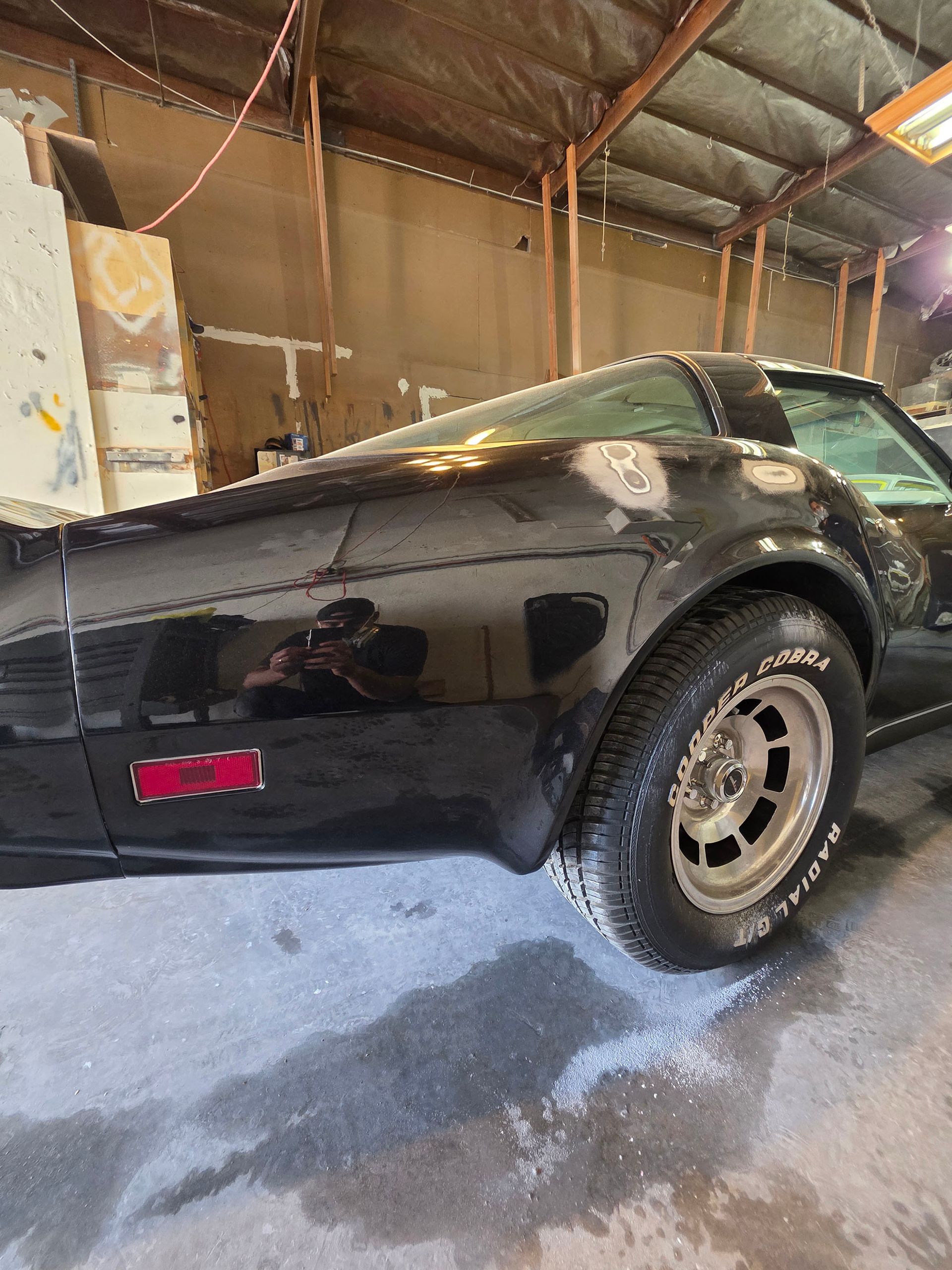 Black classic muscle car in a garage, side view with chrome wheels and wet floor.