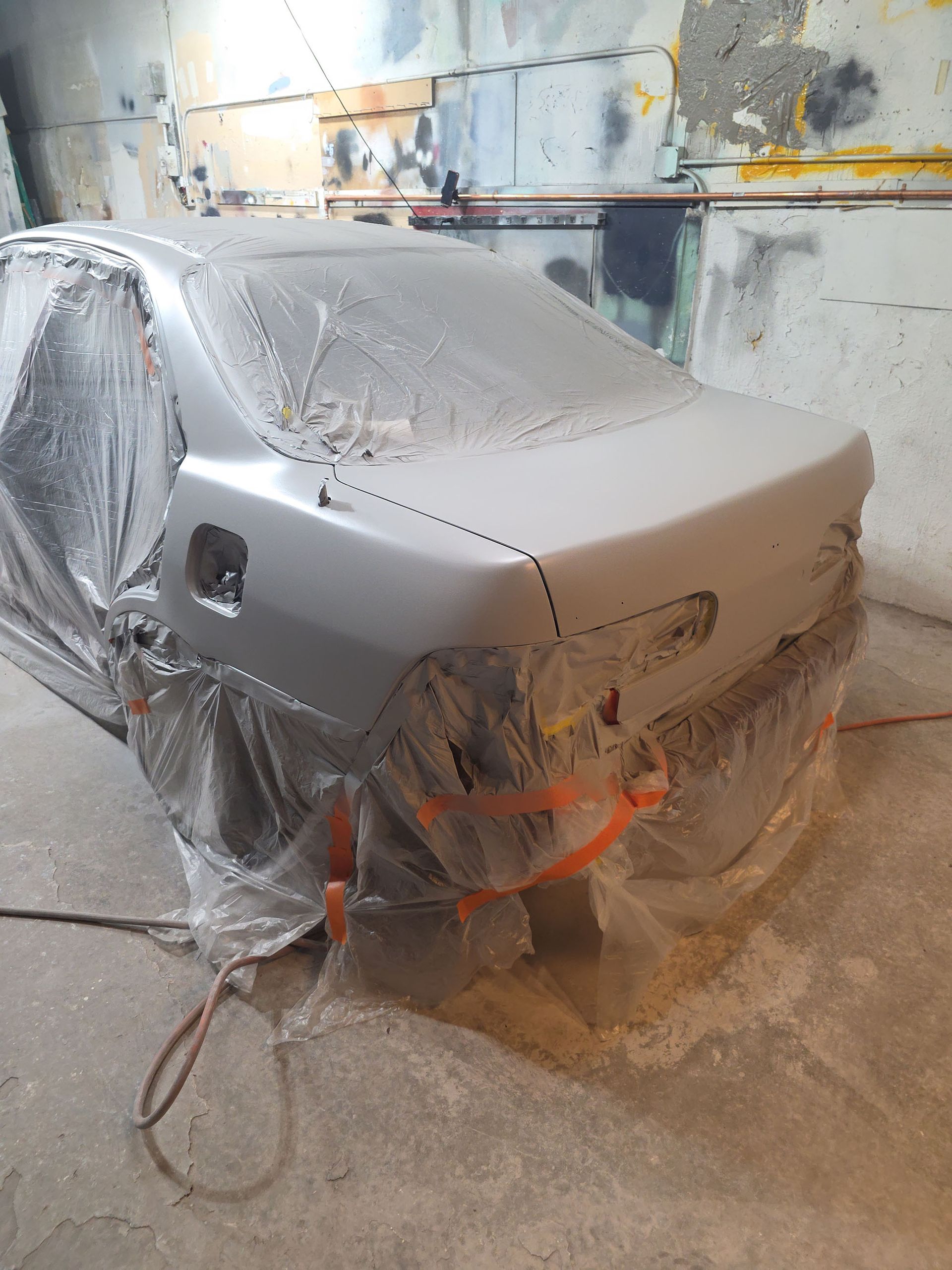 Car wrapped in plastic and primer in a paint booth, with an orange hose on the floor