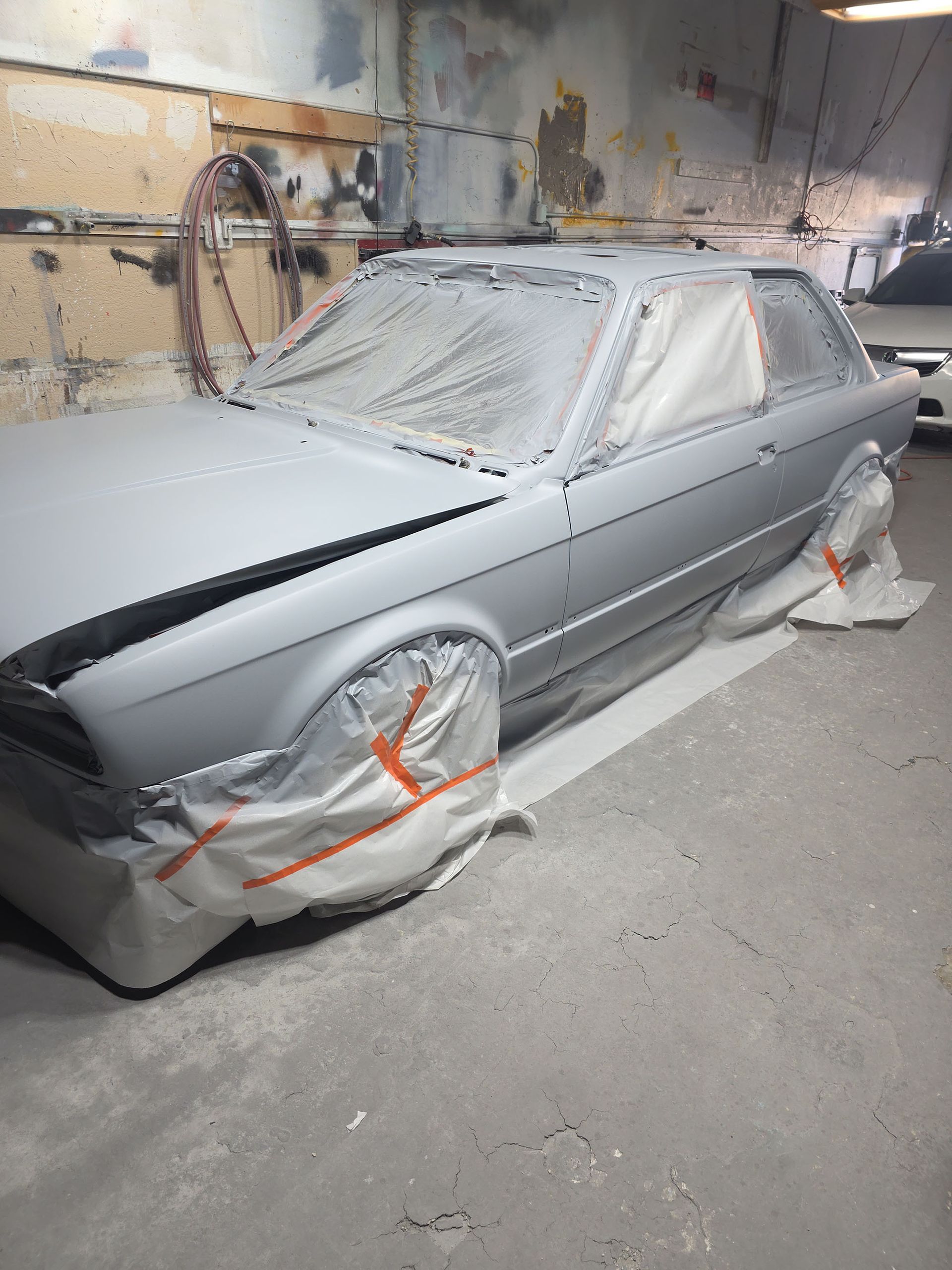 Gray sedan in a paint shop, covered in masking paper and tape for bodywork.