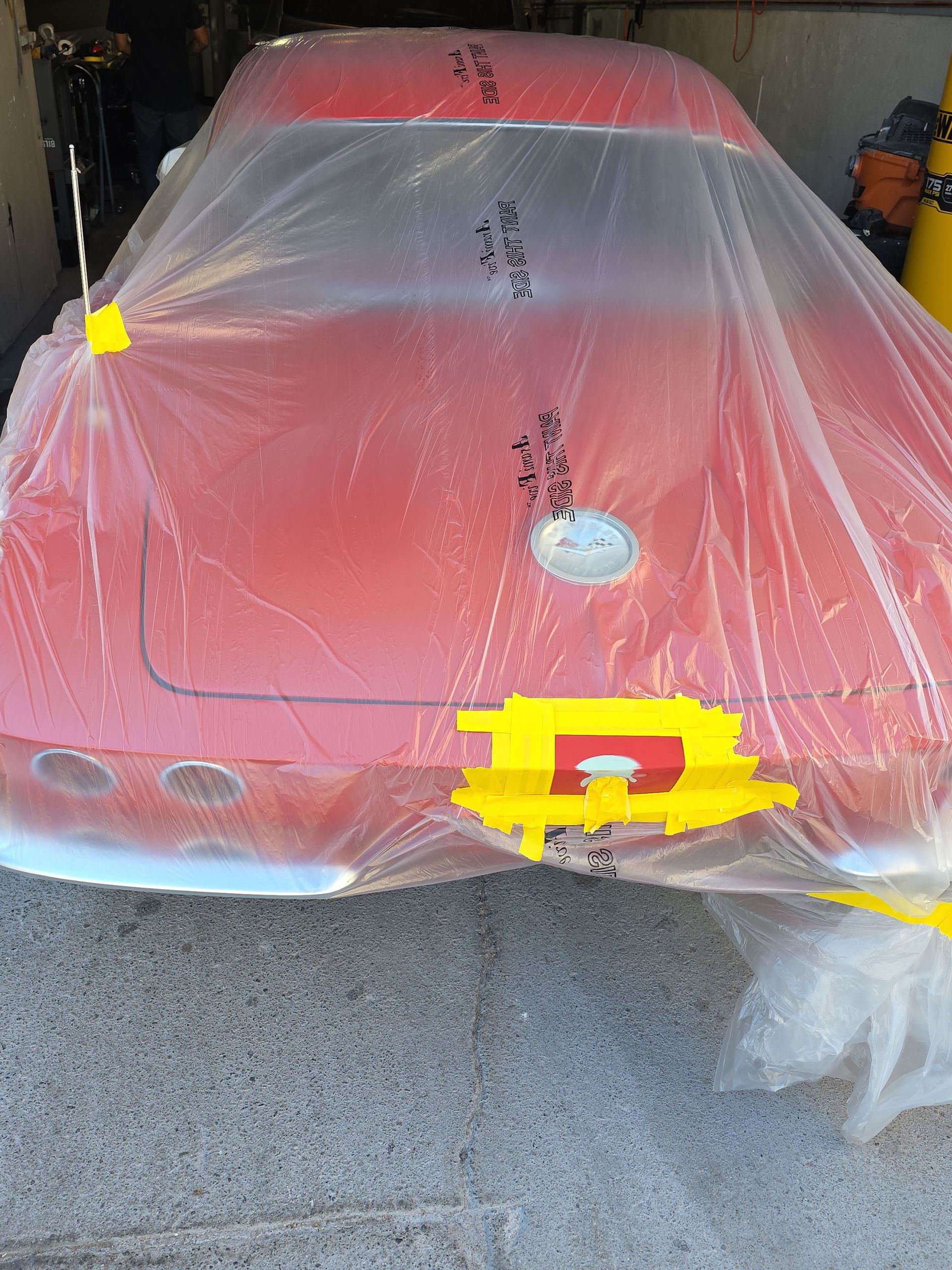 Red sports car covered in plastic sheeting inside a garage, with a yellow tape square on the front.