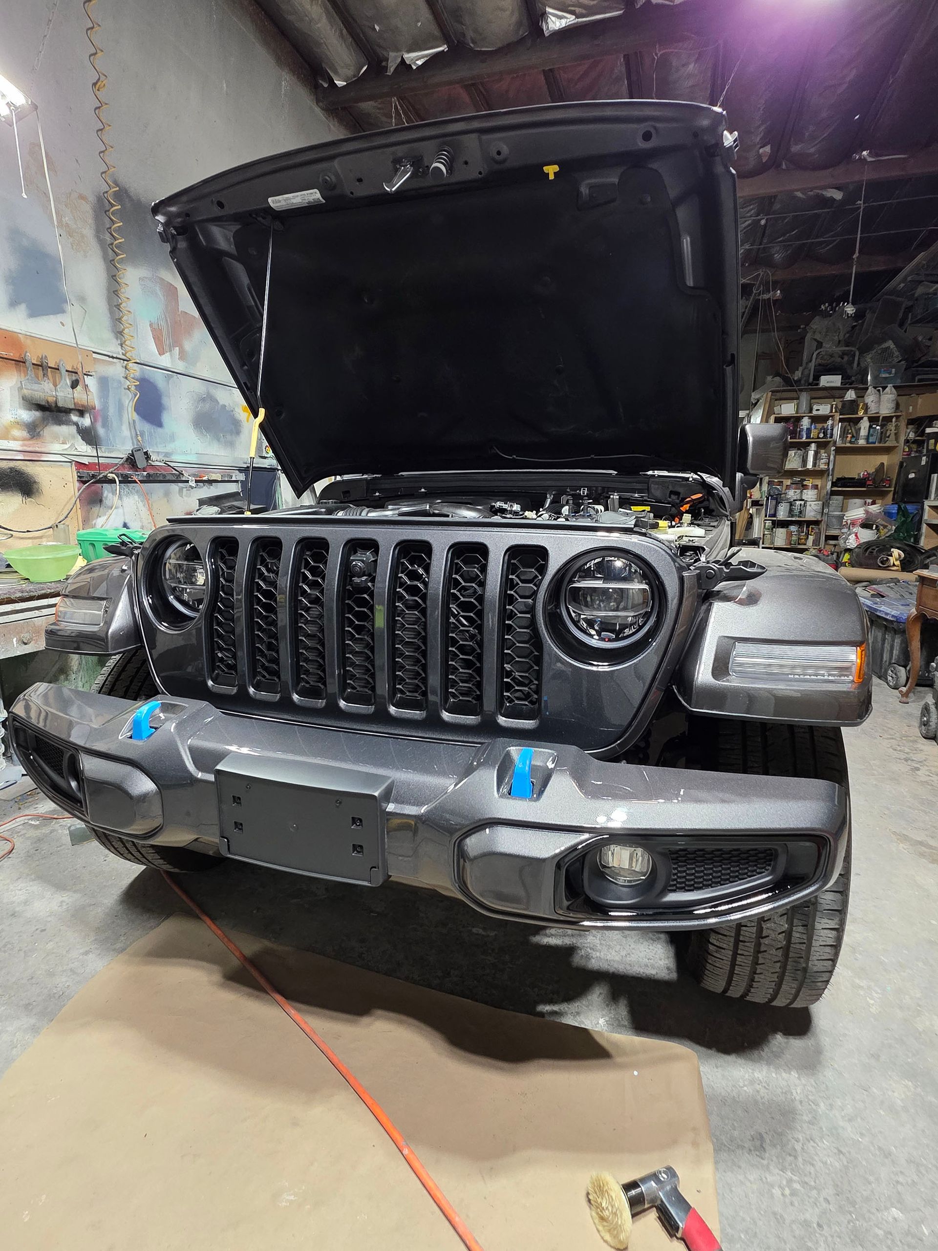 Damaged gray Jeep with raised hood in an auto repair shop, front bumper removed and headlights exposed