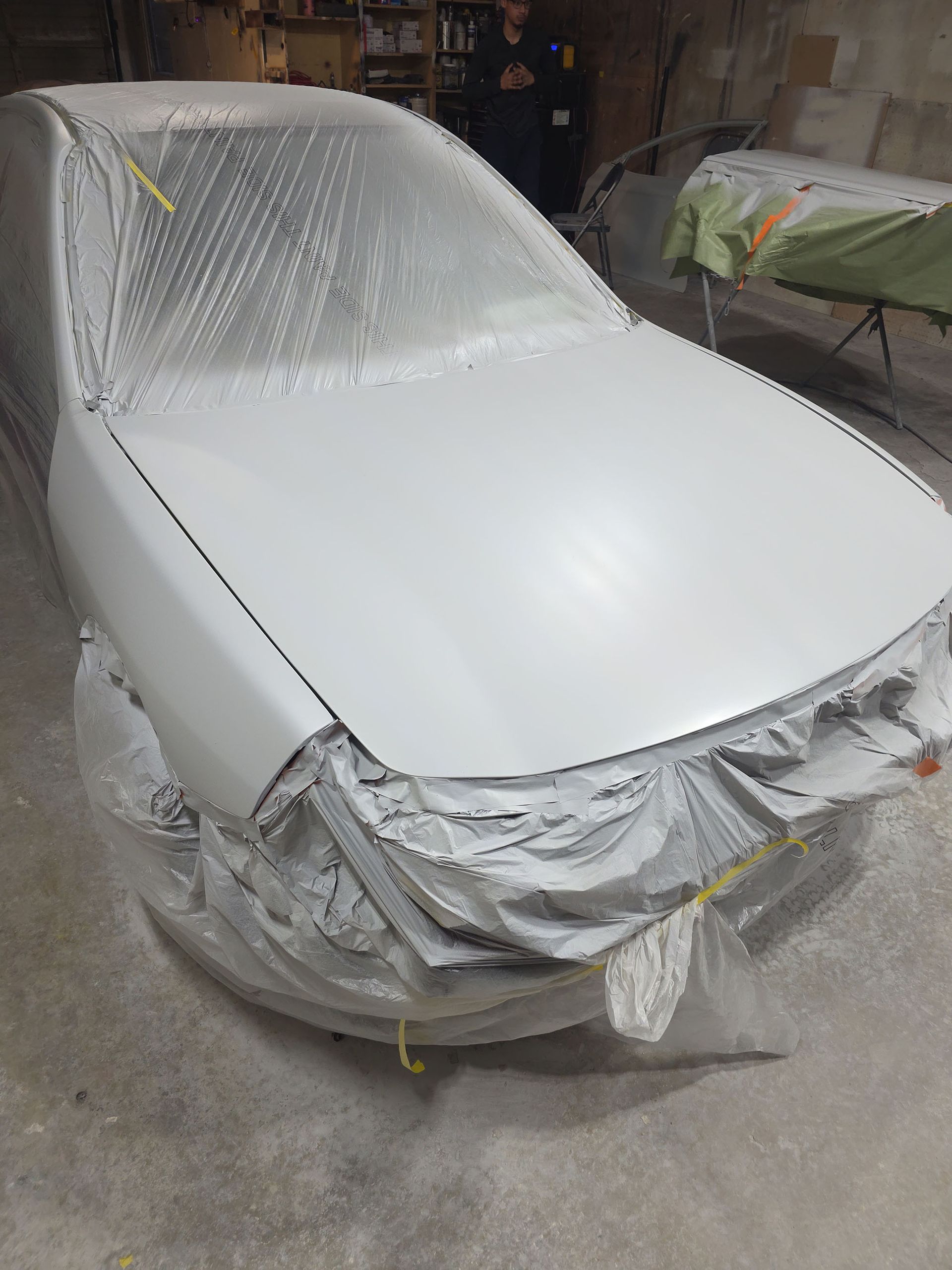 White car in a workshop, masked for painting with plastic and tape, front end partially repaired