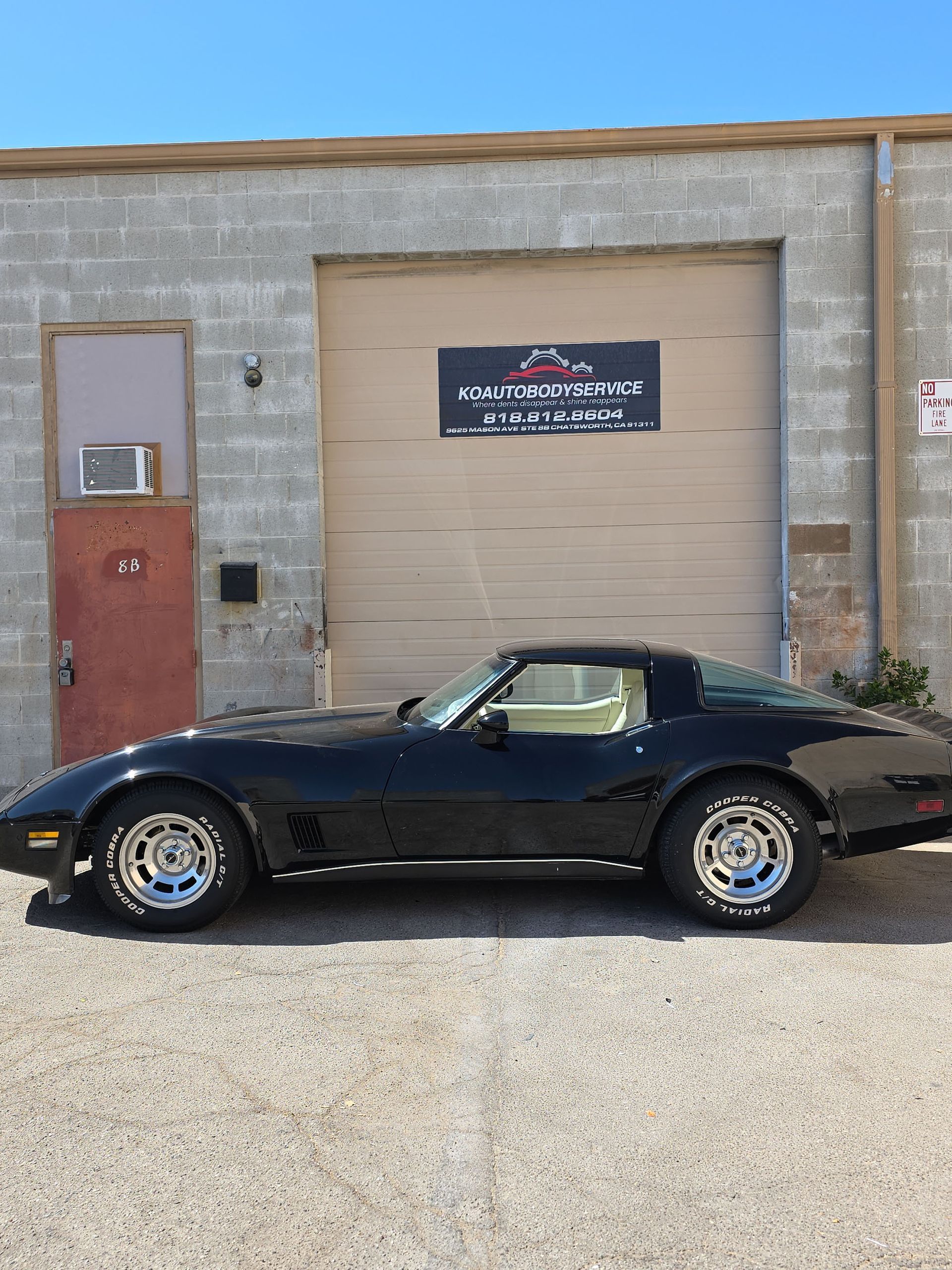 Black classic sports car parked in front of a concrete garage with a tan door and red side door