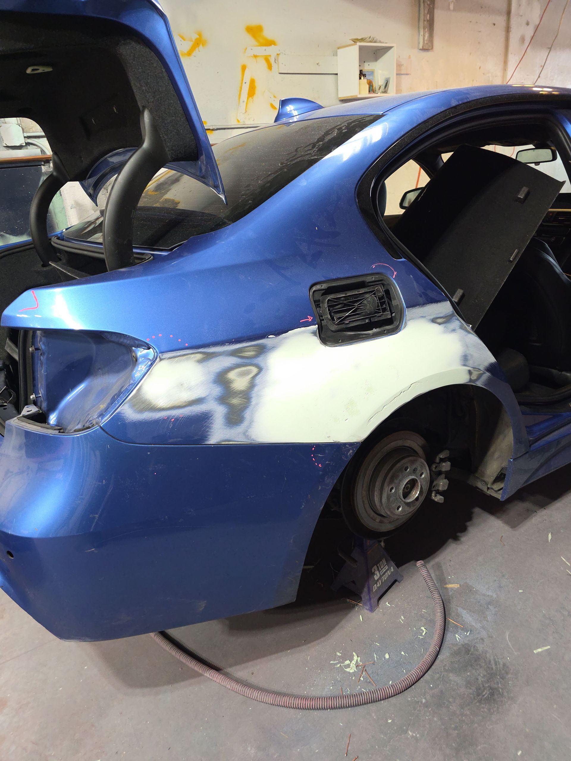 Blue compact car with side damage and missing rear wheel in a garage.