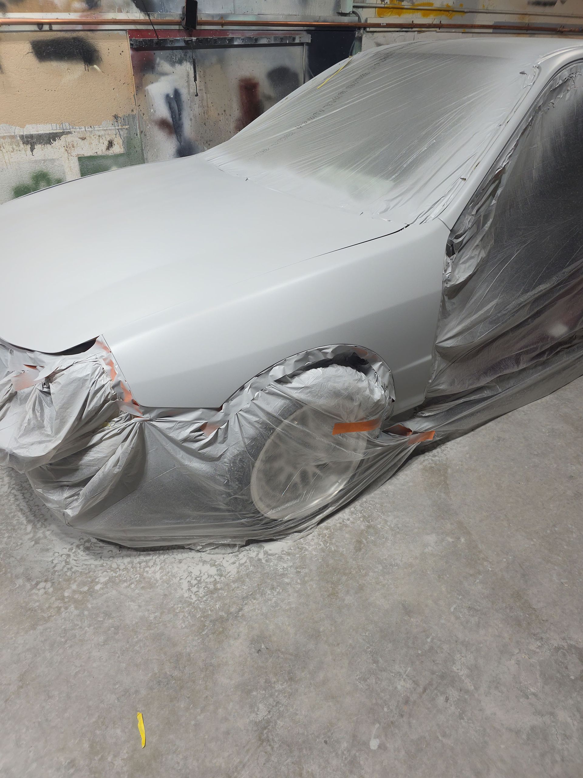 Car covered in gray primer and masking plastic in a body shop, front end visible