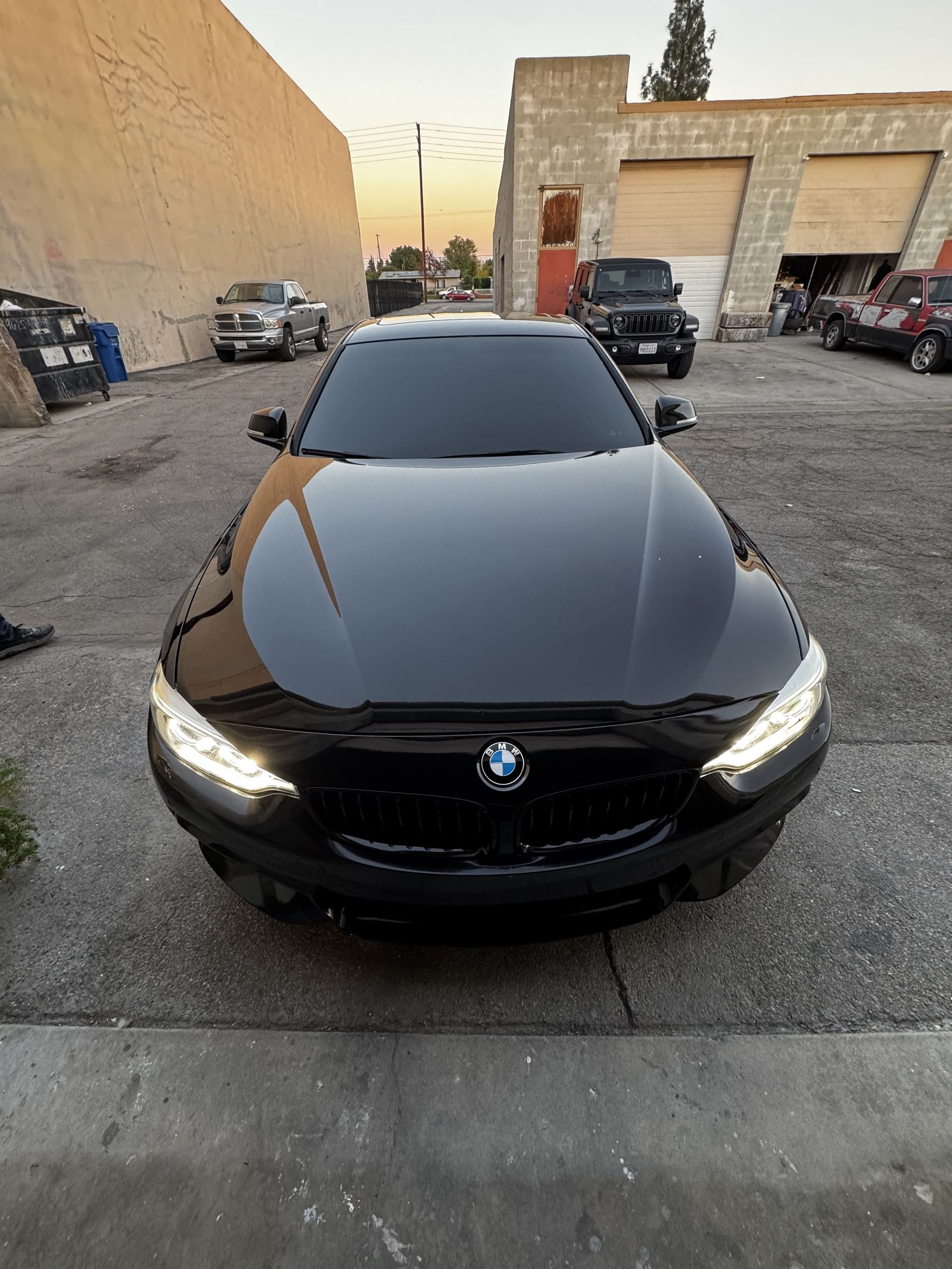 Black BMW sedan parked outdoors in a gravel lot, front view with headlights on.