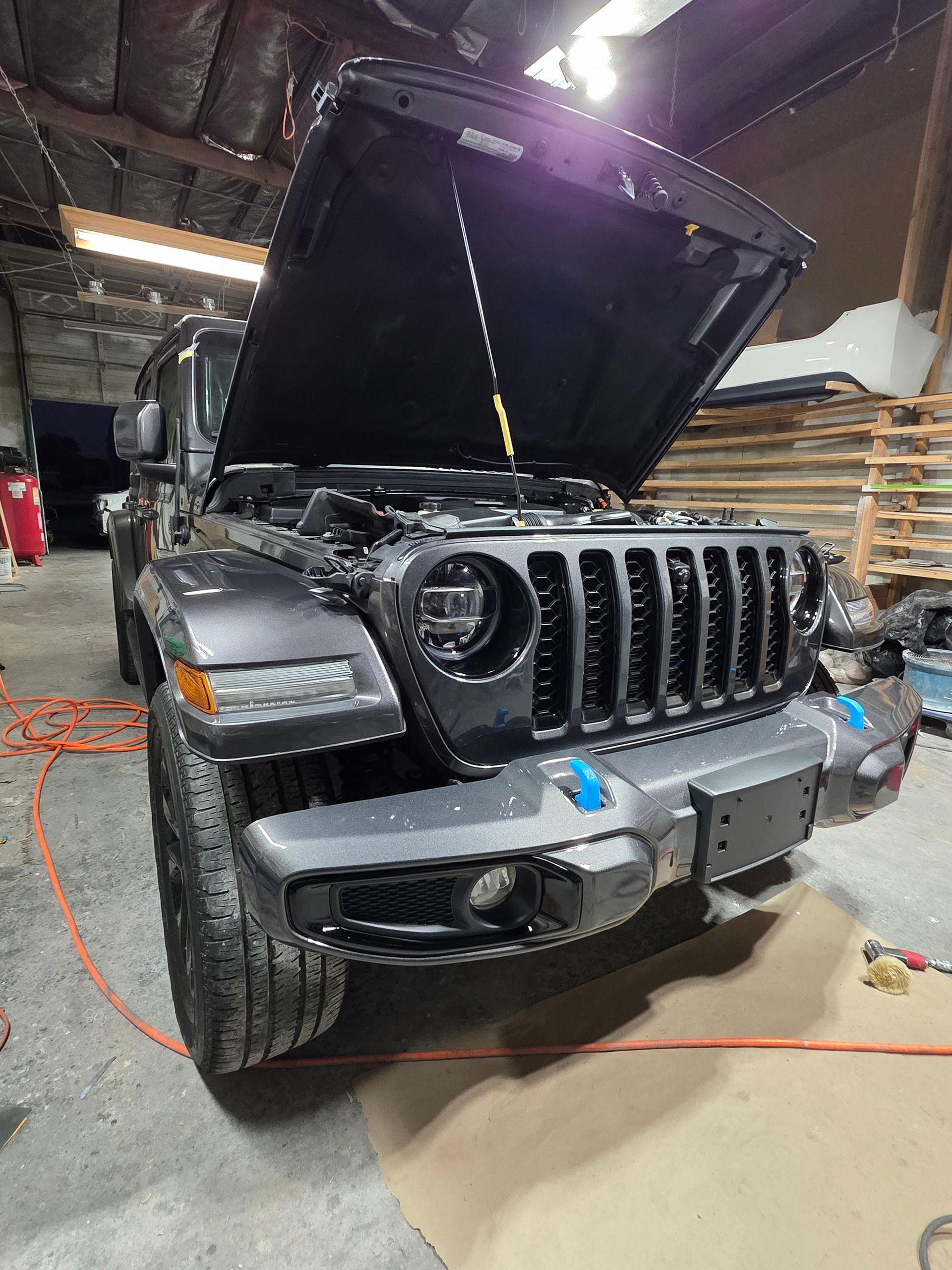 Black Jeep Wrangler with hood open inside a garage, front bumper partially removed.