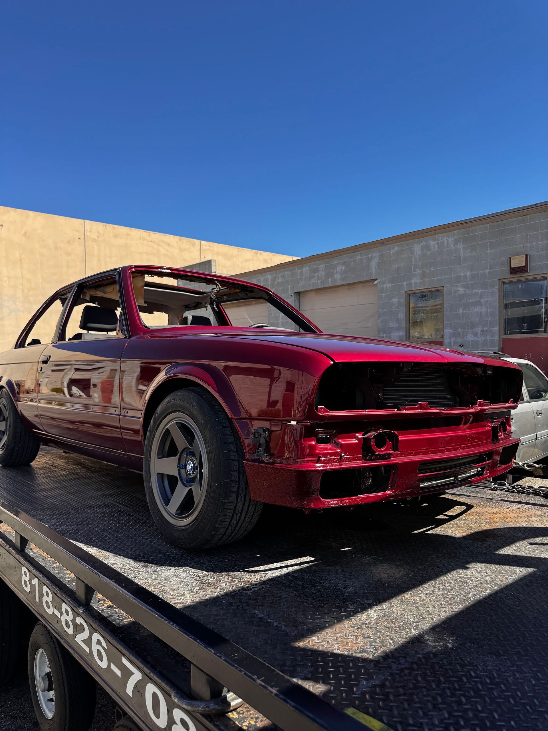 Damaged red race car on a flatbed trailer outside a warehouse under a blue sky
