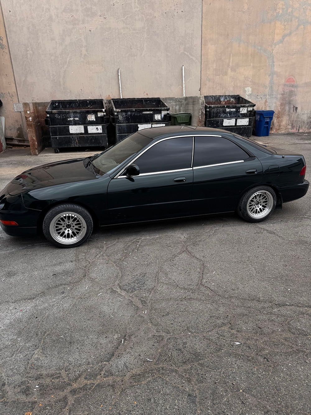 Black two-door coupe parked beside dumpsters against a beige wall.