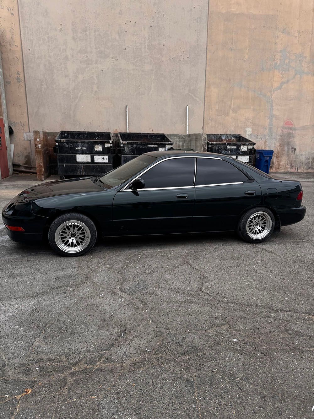 Black sedan parked beside a concrete wall and dumpsters on a gravel lot.