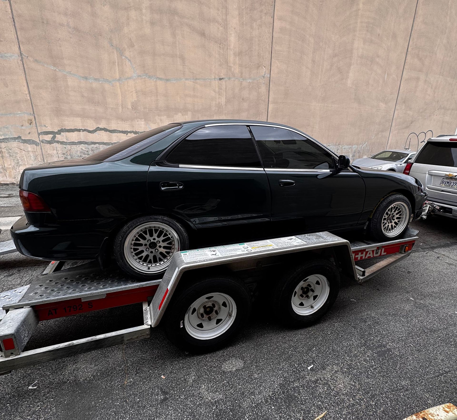 Black sedan strapped on a twin-axle trailer, parked beside a beige wall