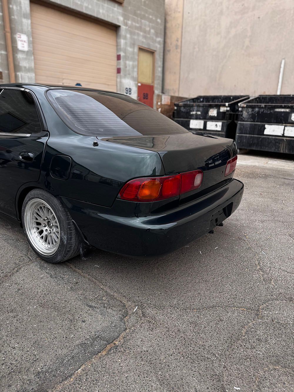Black sedan parked in an alley beside dumpsters and a tan building