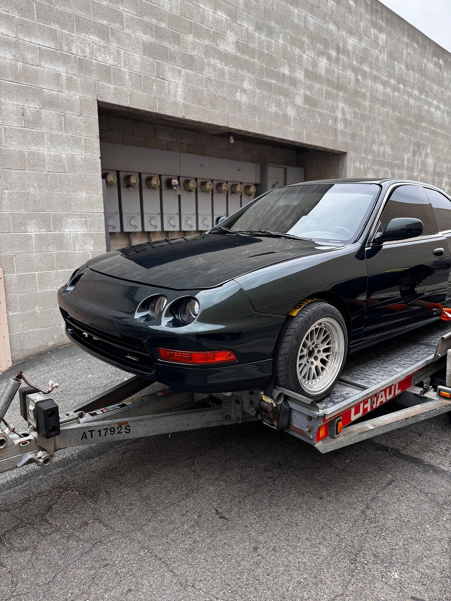 Black hatchback sports car secured on a trailer beside a gray cinderblock wall.