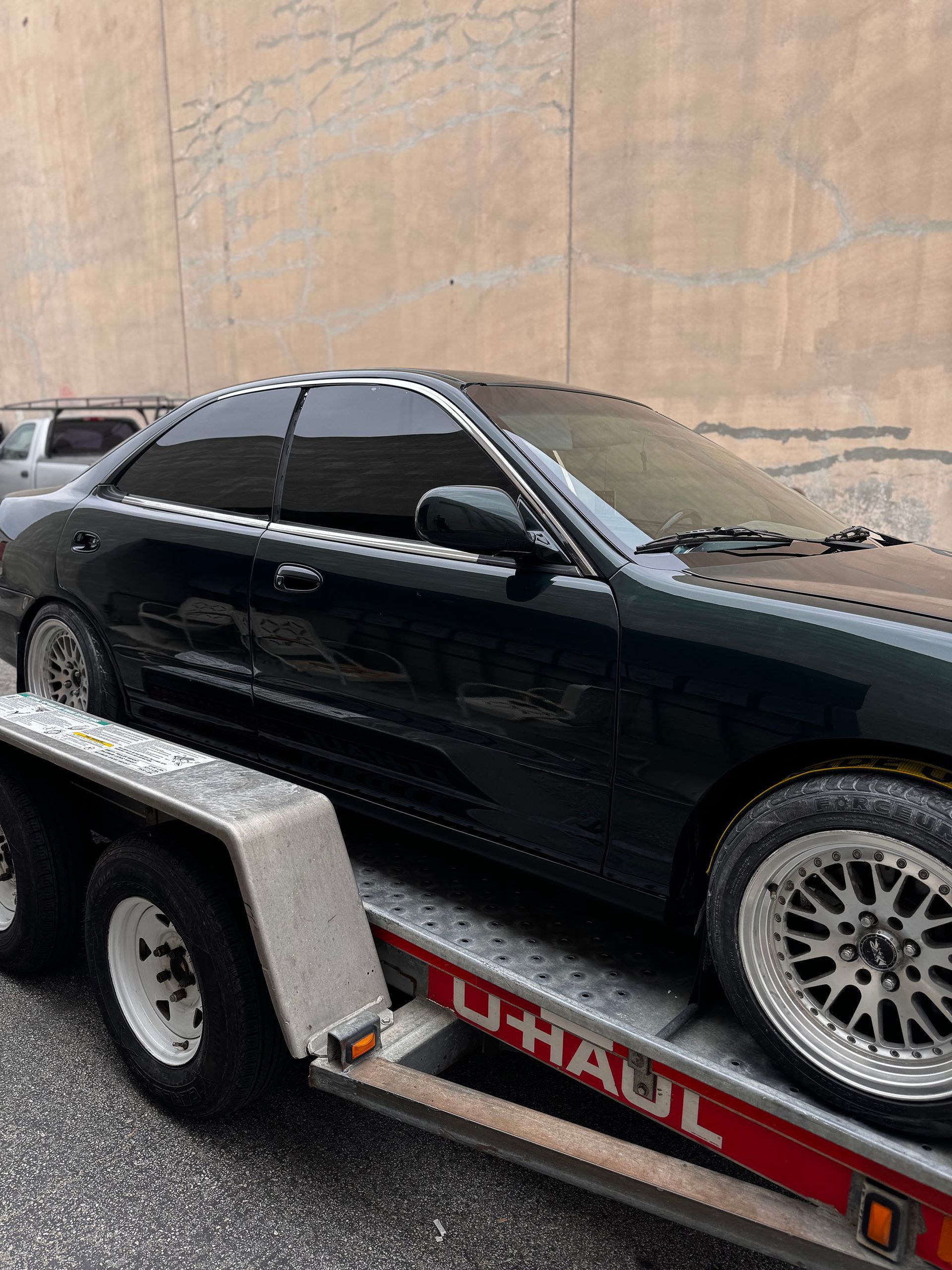 Black coupe on a U-Haul trailer beside a tan wall, with tinted windows and silver rims