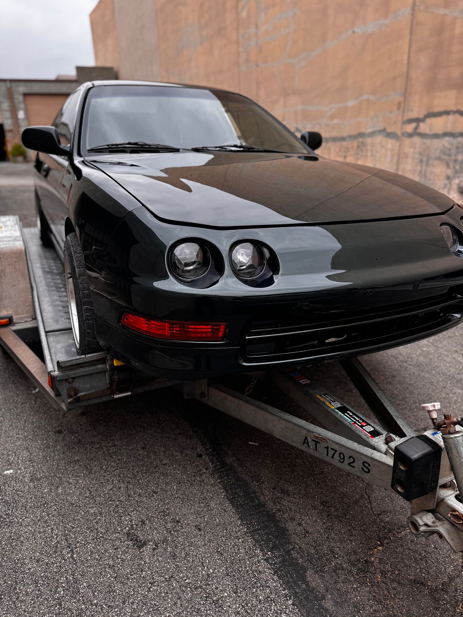 Black sports car secured on a flatbed trailer on a wet street, with its front bumper and headlights visible.