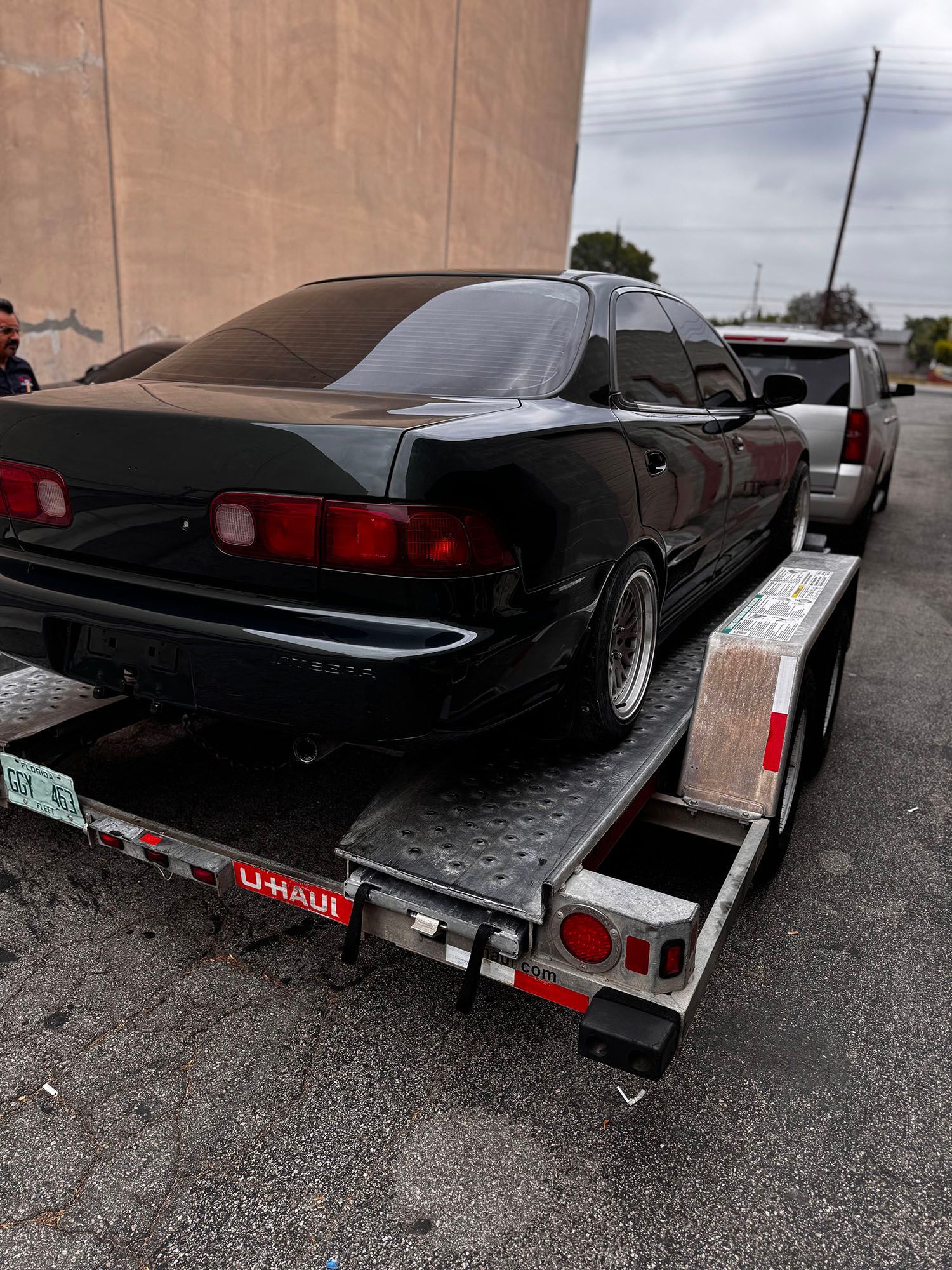 Black sedan on a car trailer in a gravel lot, rear view, with a white SUV behind it.