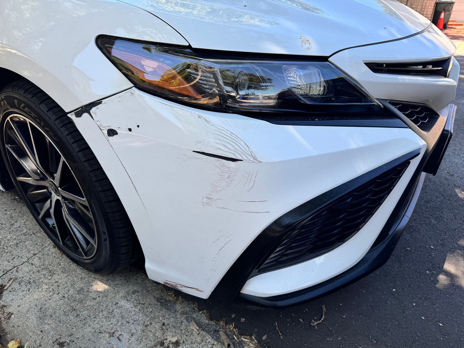 Close-up of a white car’s front bumper and headlight with scrape marks on the left side.