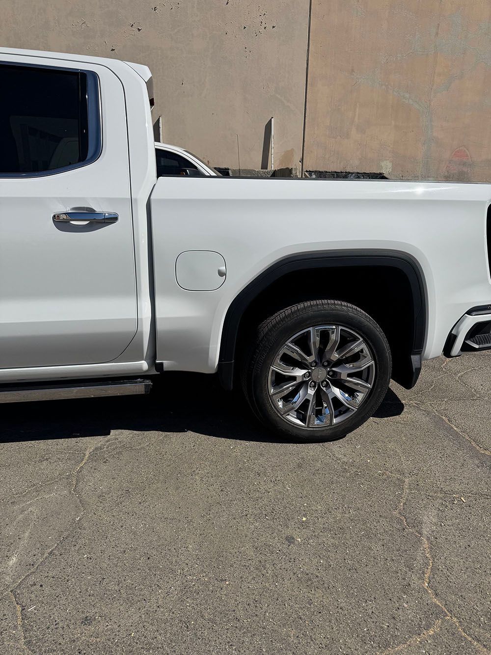 White pickup truck parked beside a beige wall on gravel, showing the rear side and wheel.