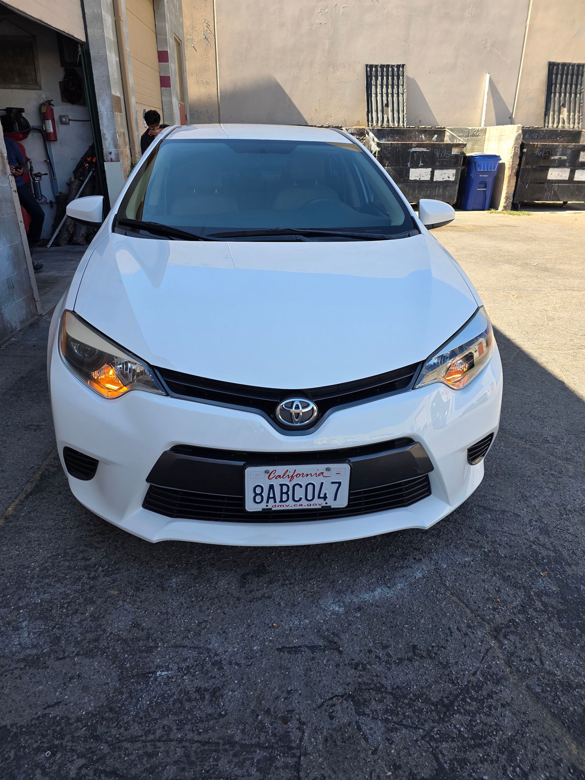 White Toyota sedan parked on gravel in front of a building, headlights on.