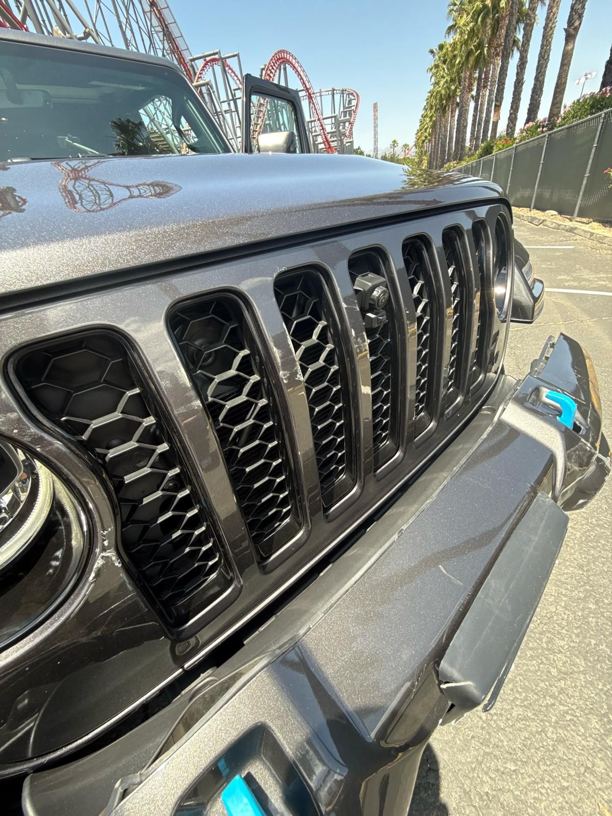 Close-up of a gray Jeep grille and front bumper on a sunny street, with a roller coaster in the background