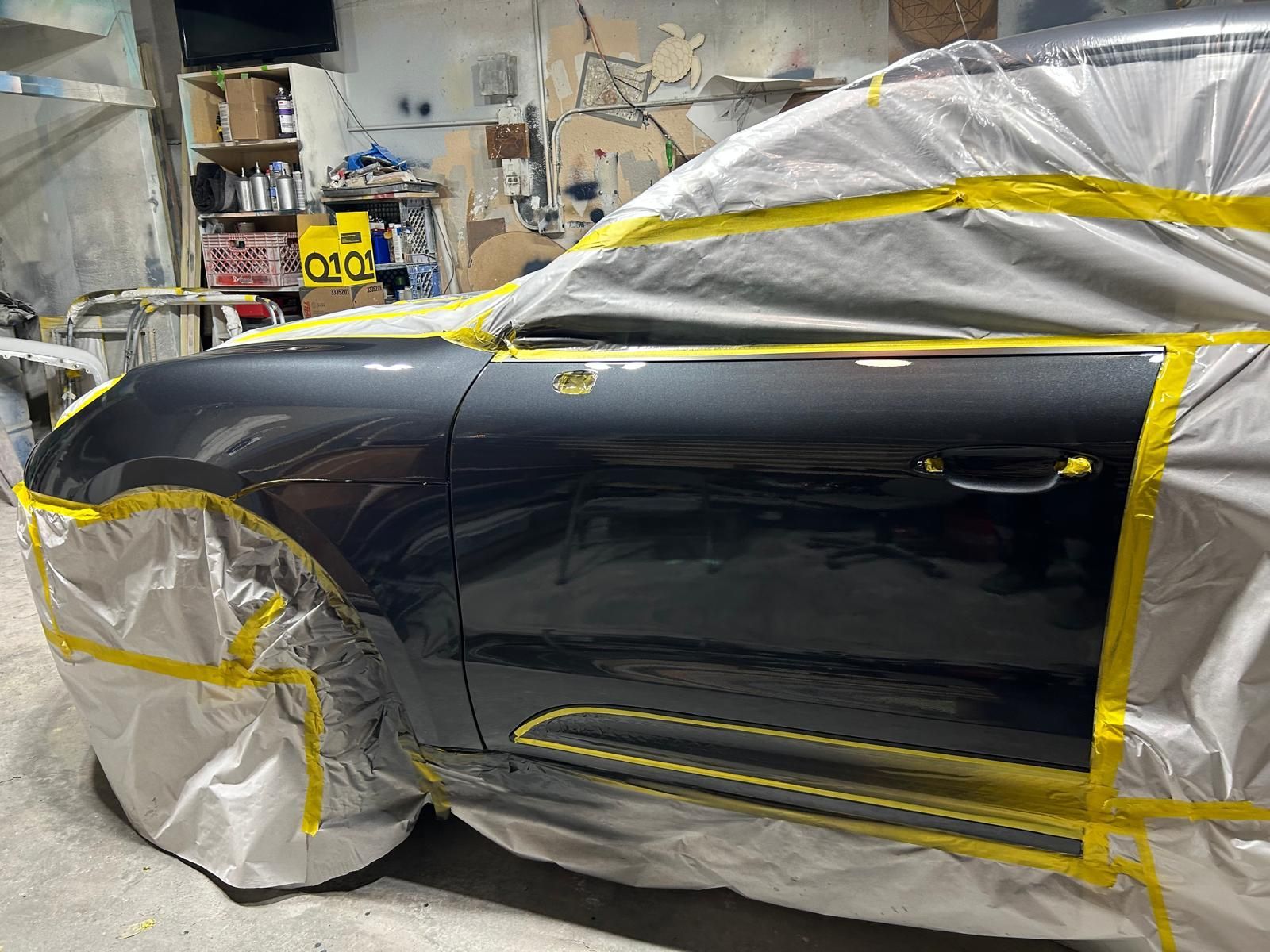 Black car in a body shop, masked with plastic and tape during paint preparation.