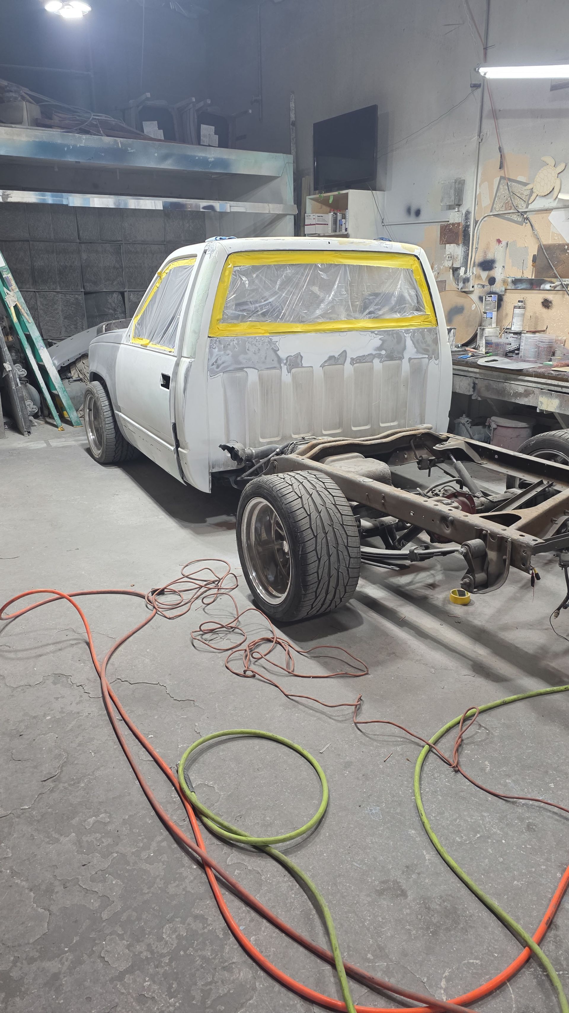 Primered pickup truck chassis in an auto shop, with yellow tape on windows and tools on the floor.