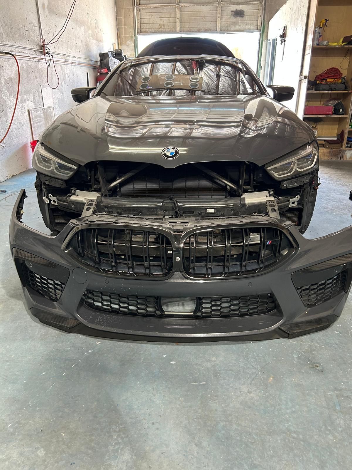 Black BMW front end heavily damaged in a garage, with the bumper detached and grille exposed