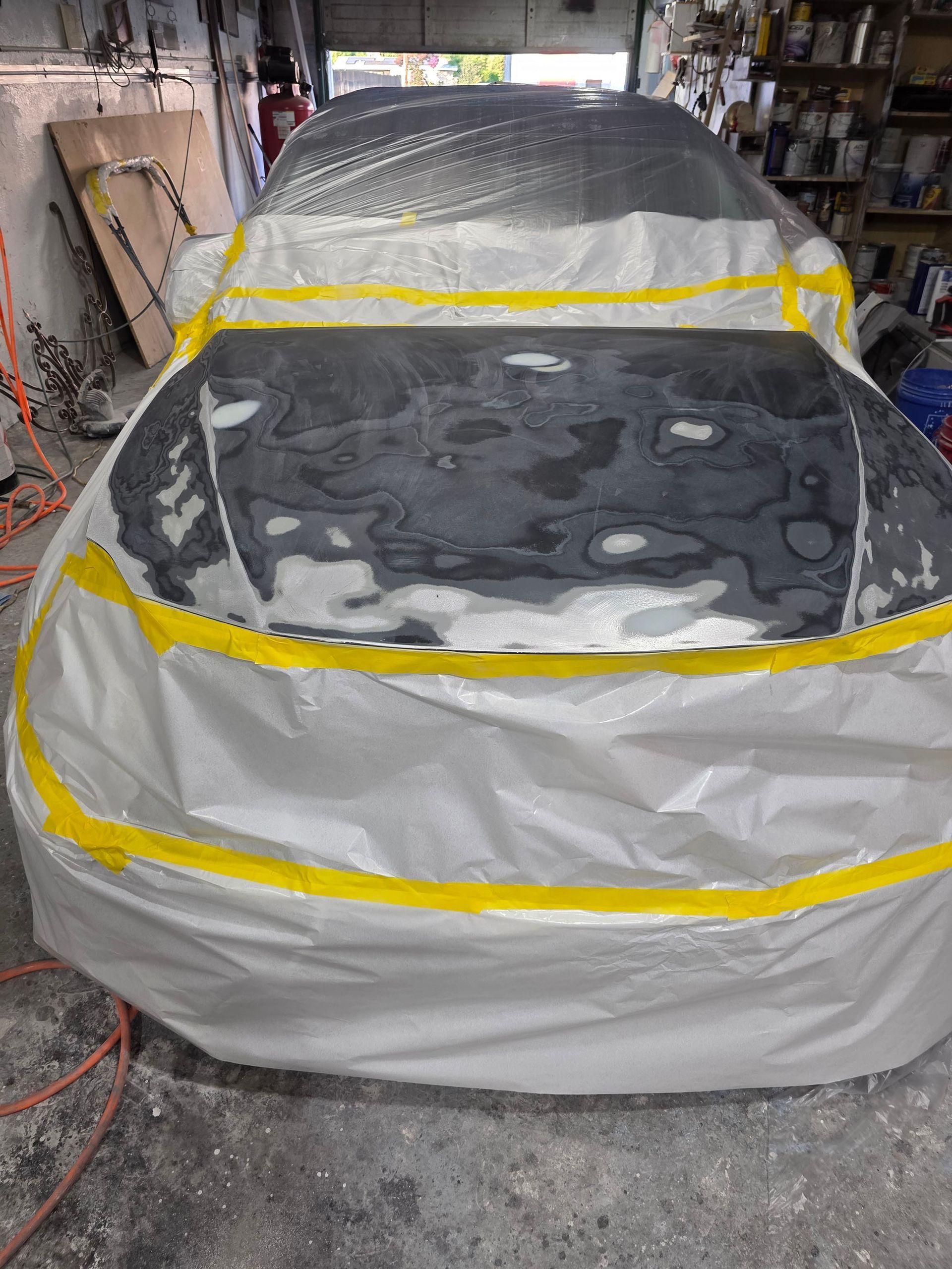 Car covered in plastic and tape in a cluttered garage, likely being painted.