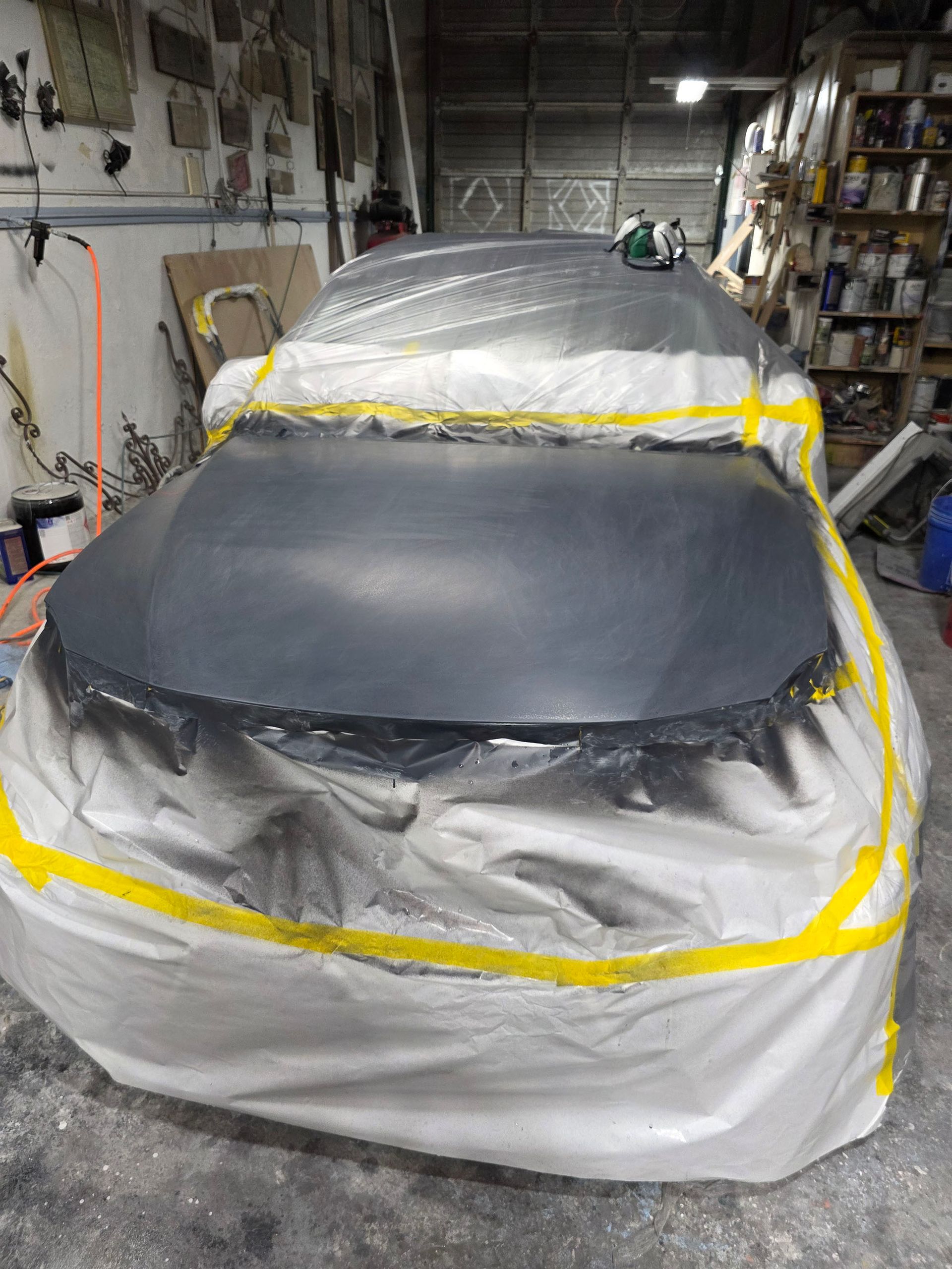 Car covered in masking paper and tape inside a garage, with the hood and front end primed black.