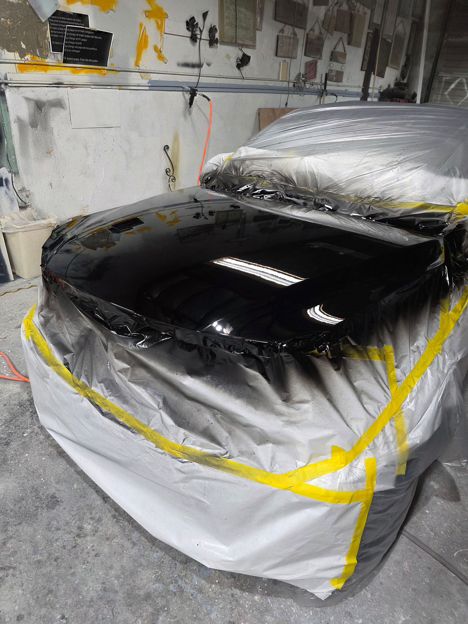 A black vehicle hood being spray-painted in a masked-off auto body shop booth with yellow tape and plastic sheeting