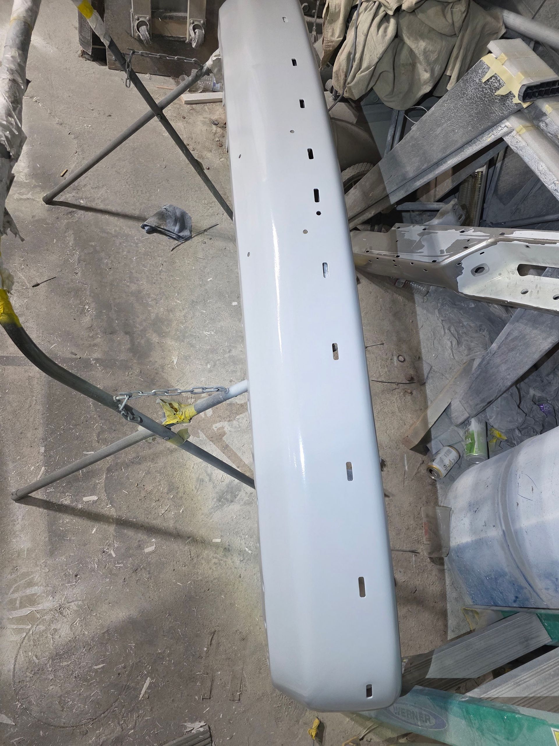 White car bumper lying on a concrete floor in a workshop, with tools and parts nearby