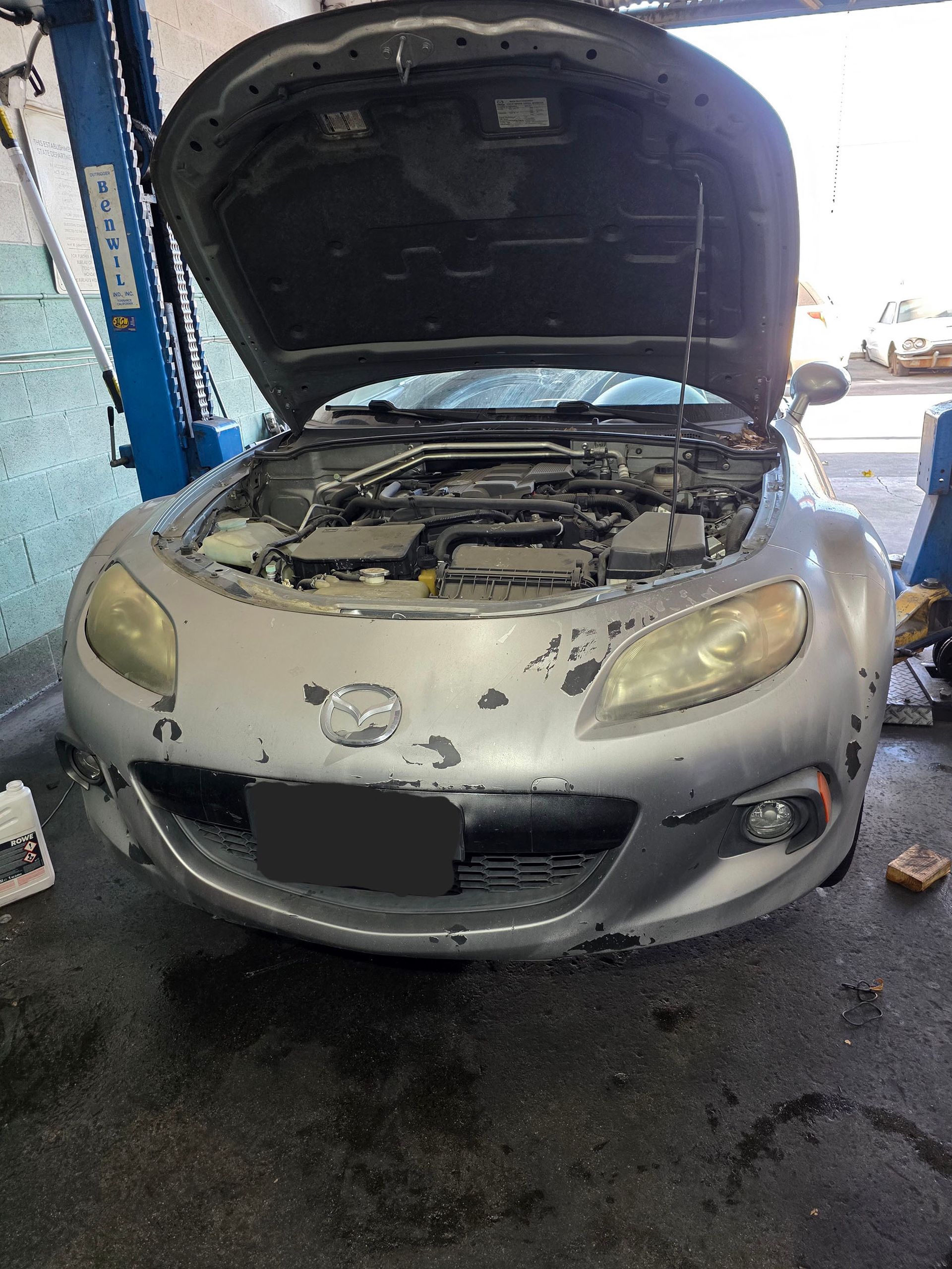 Damaged silver car with hood open in a repair garage, front bumper and headlights scratched.